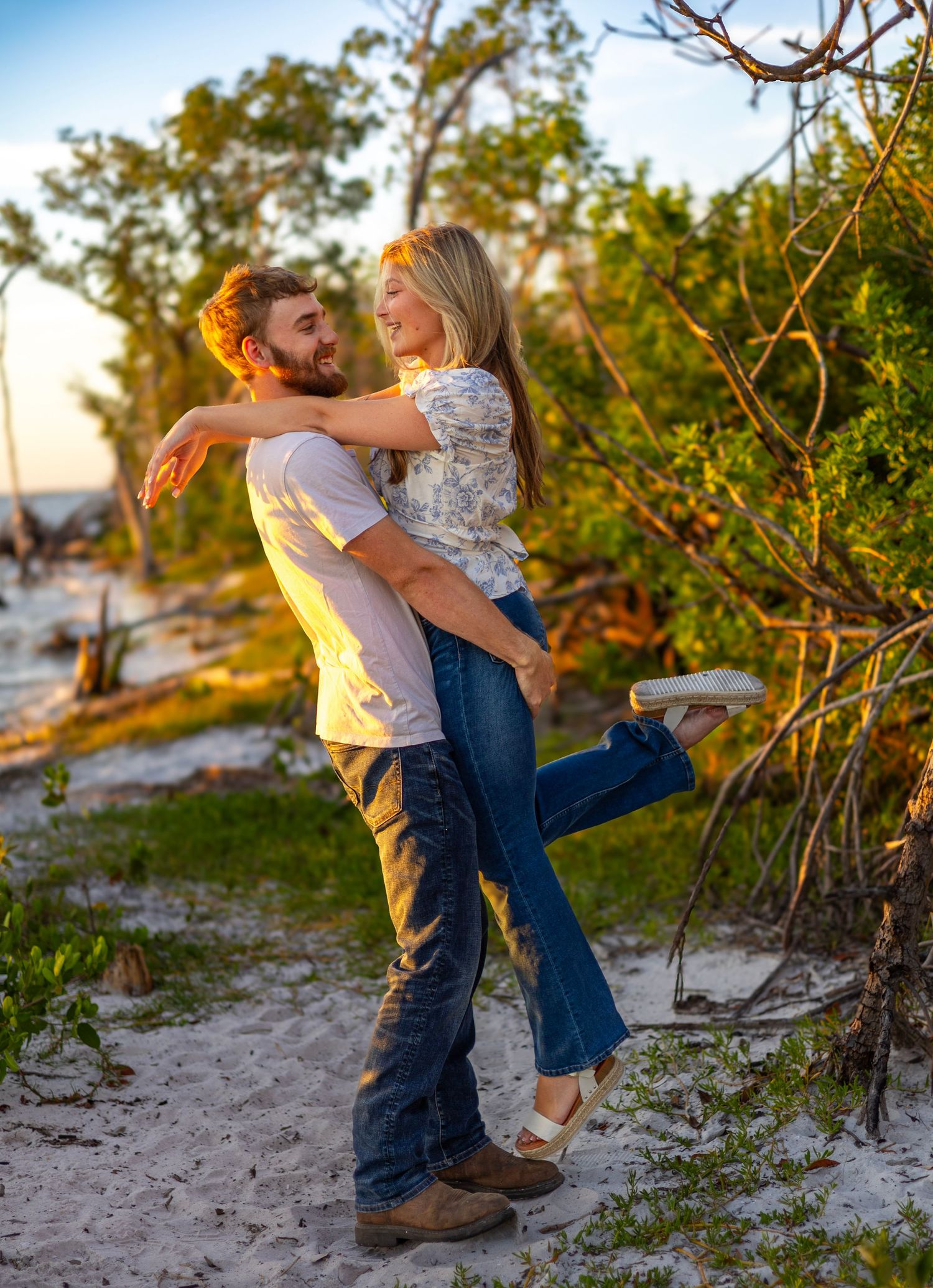A couple embraces joyfully on a sandy beach at sunset, surrounded by lush green trees in a romantic outdoor setting.