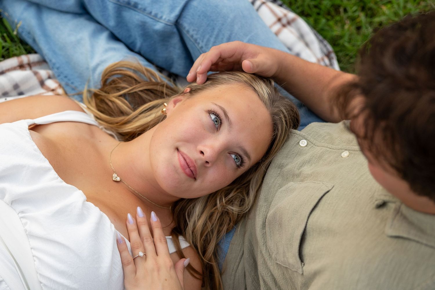 A young woman with blonde hair lies on grass while a man gently touches her head during a romantic outdoor moment.