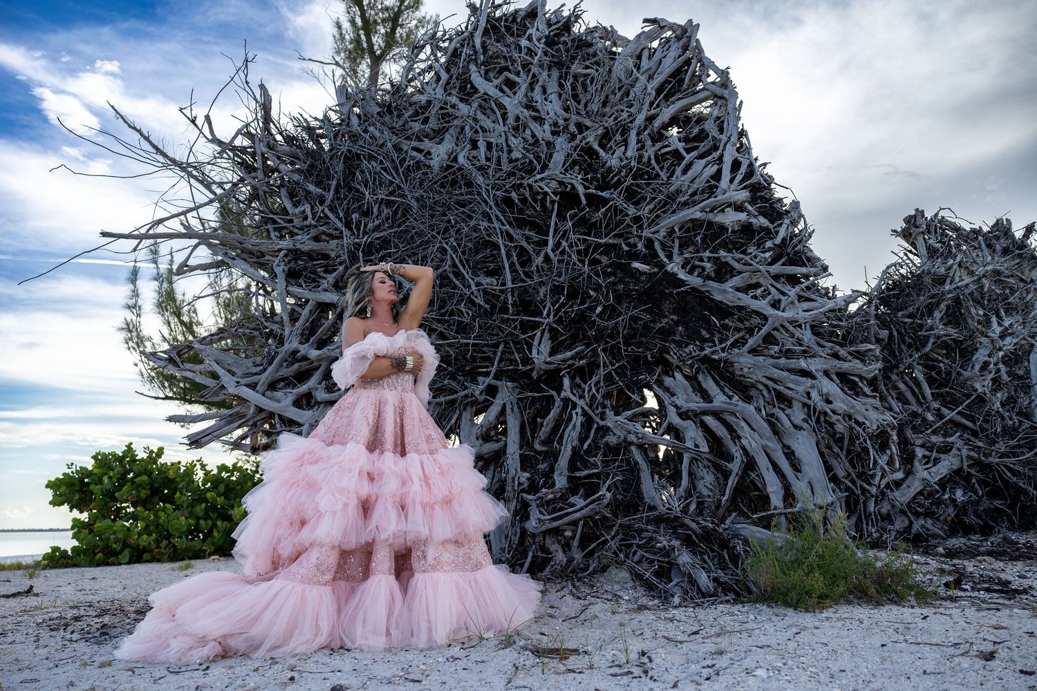 Woman in pink tiered ball gown poses on sandy beach before dramatic driftwood sculpture under cloudy sky.
