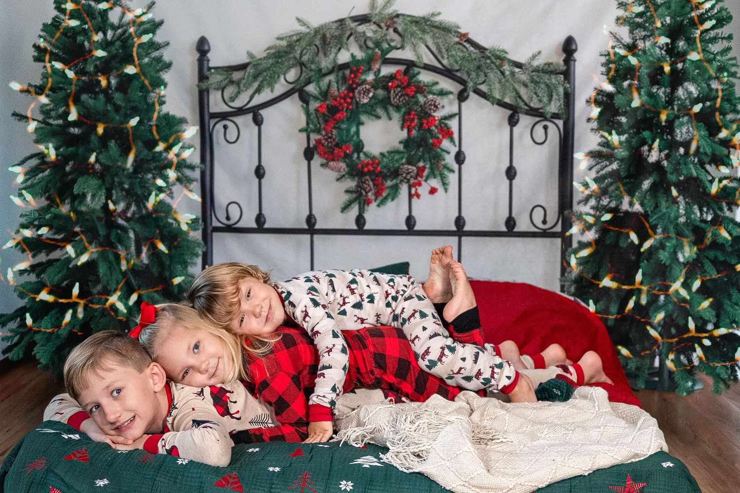 Three children in Christmas pajamas lying on a bed surrounded by decorated holiday trees and a festive wreath backdrop.