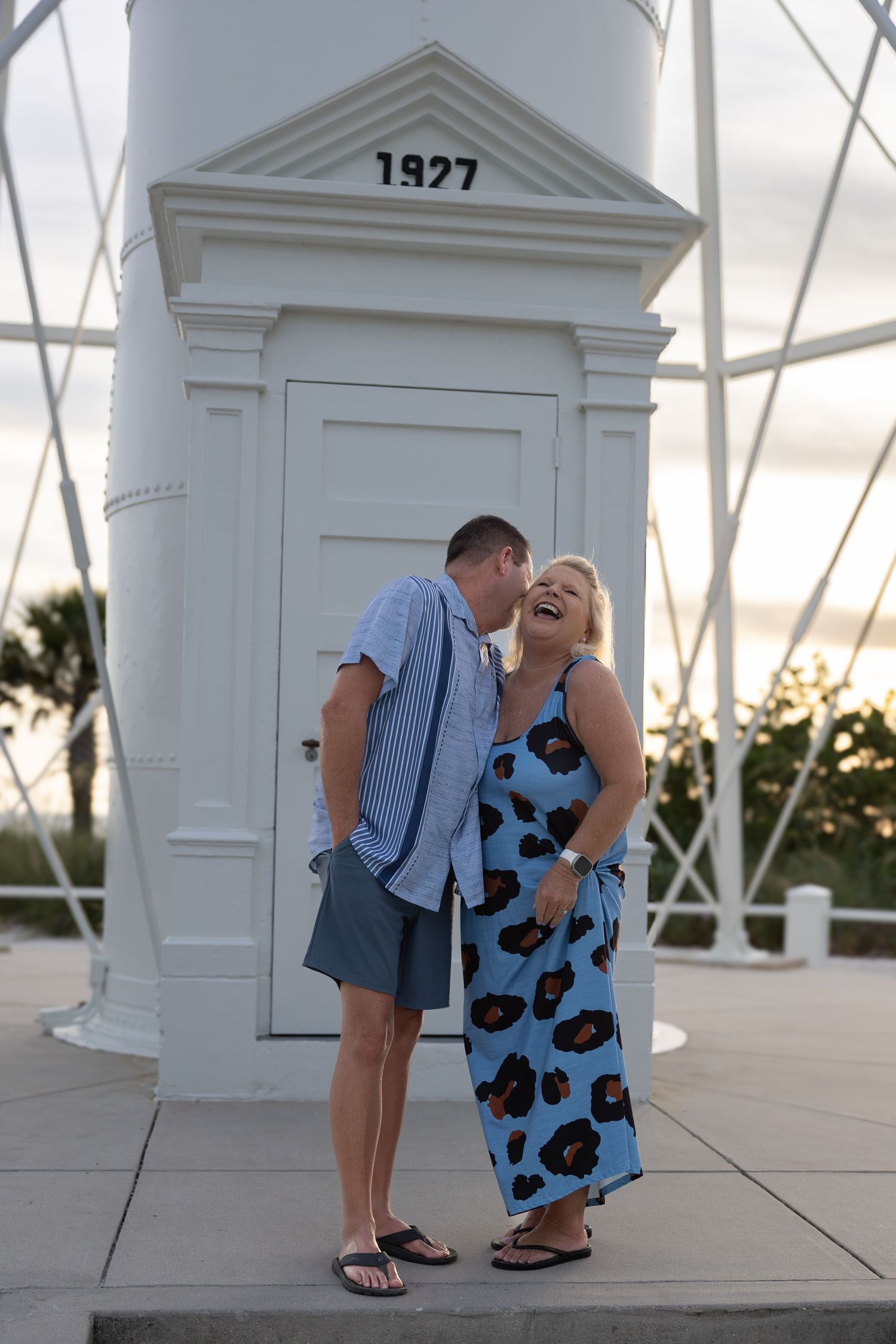 Couple laughing together in front of a white 1927 lighthouse at sunset, wearing casual summer outfits.