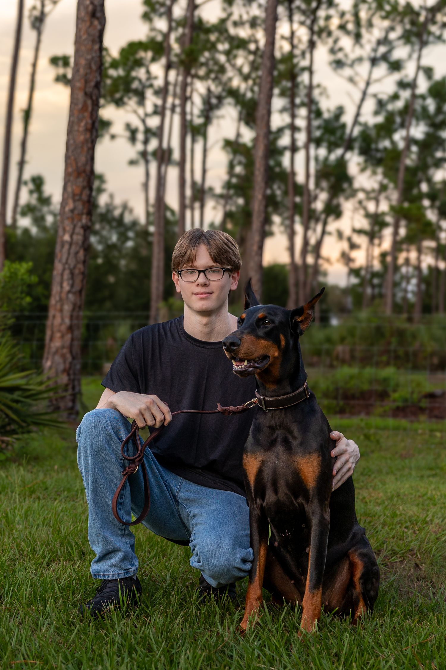 Young man with glasses in black tee kneeling beside a Doberman Pinscher on a leash in a lush green yard.