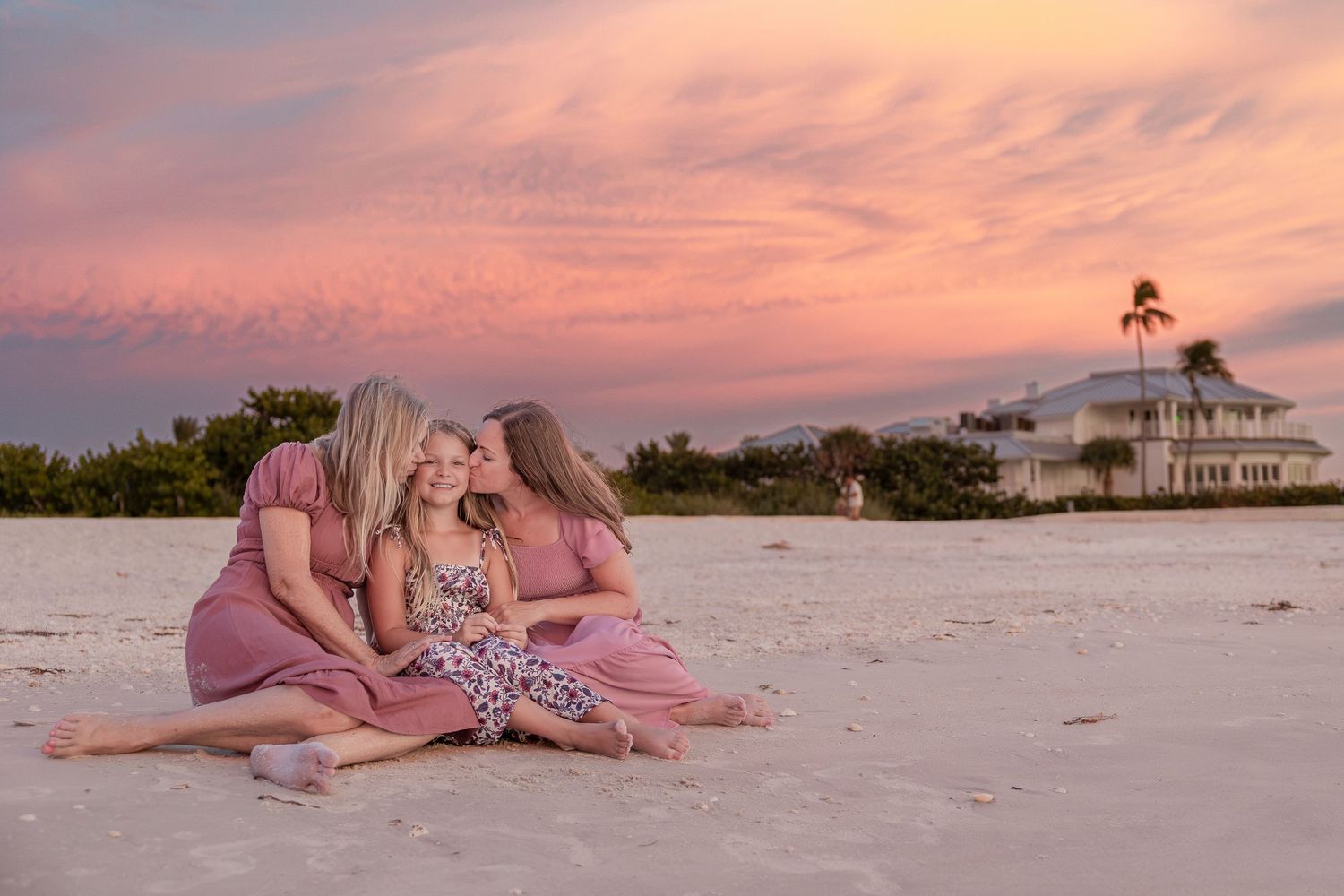 Three women in pink dresses sit together on a white sand beach at sunset with a coastal house in the background.