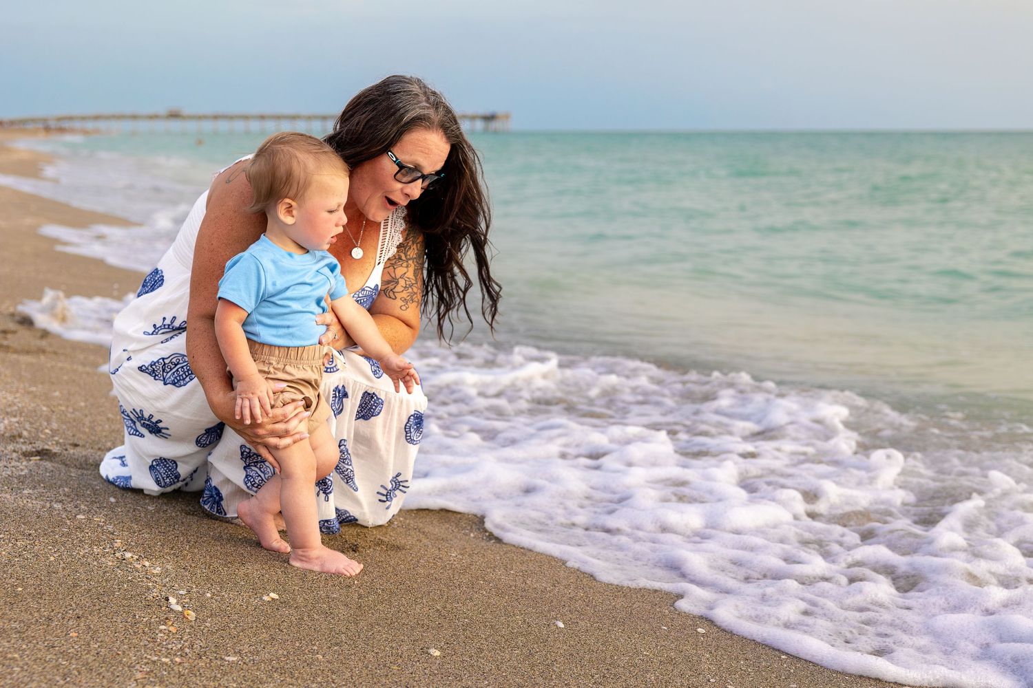 Mother and baby boy sitting together on a sandy beach shore, watching ocean waves on a sunny day.