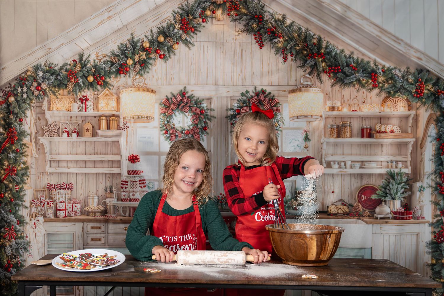 Two girls bake Christmas cookies in festive holiday kitchen backdrop with wreaths and garland decorations.