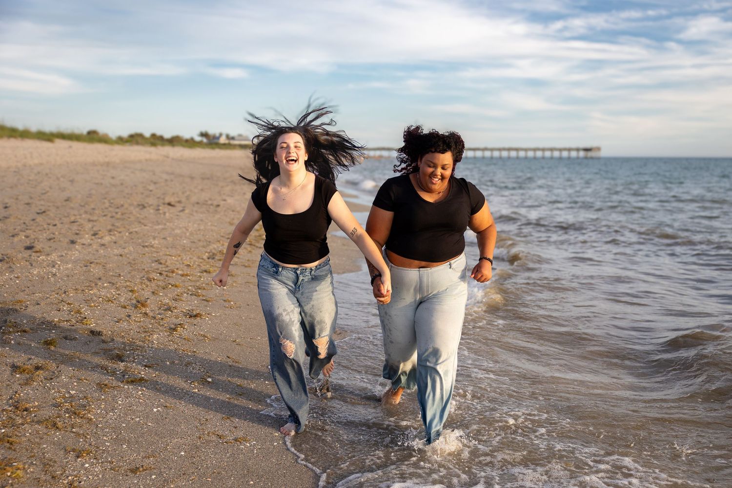 Two joyful women running hand-in-hand through shallow ocean waves on beach.
