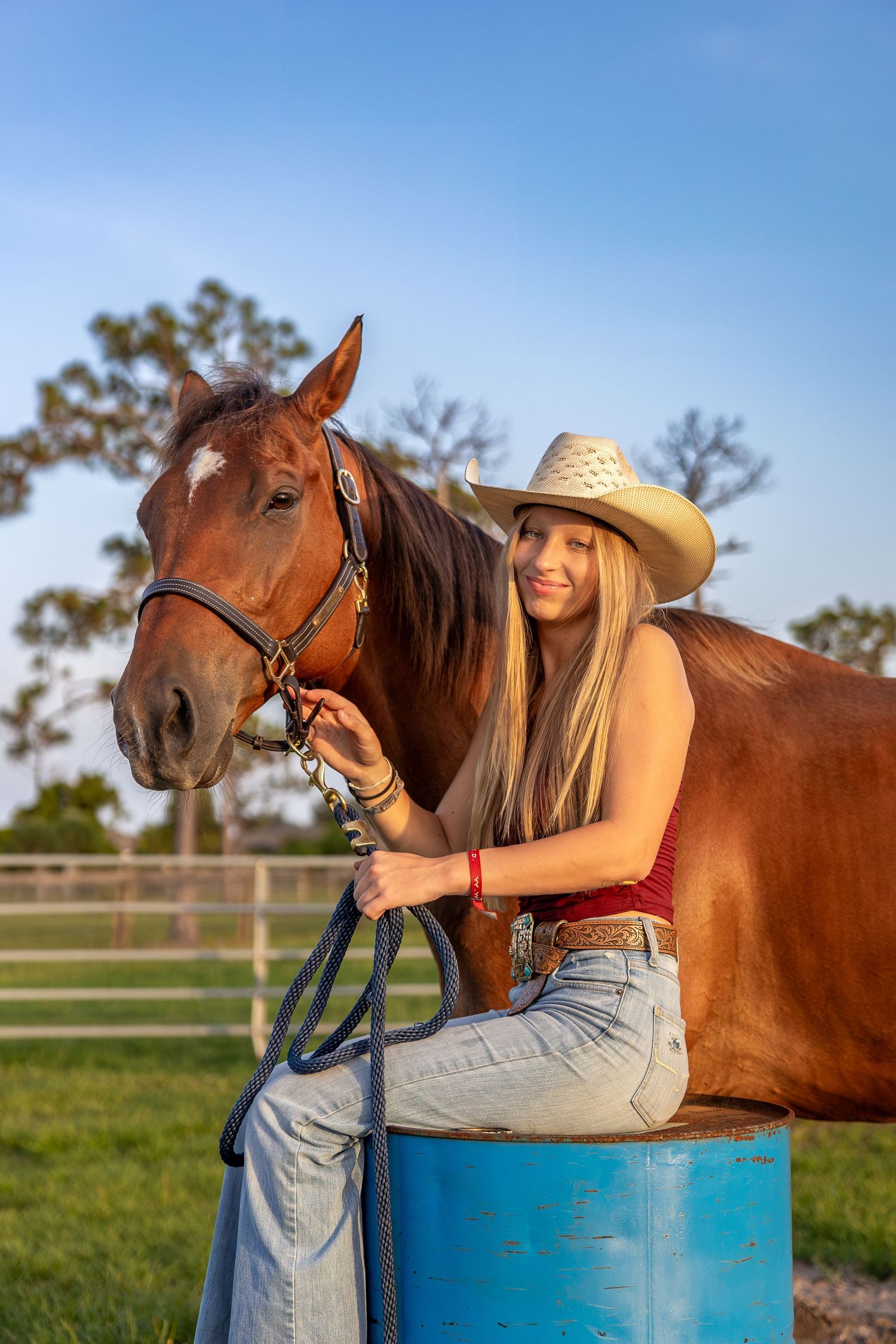 Young blonde woman in cowboy hat and jeans sits on barrel beside chestnut horse in sunny ranch setting. Senior photo session.