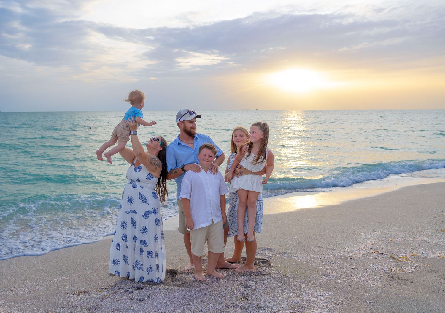 Family of six poses on a sandy beach at sunset with turquoise ocean waves, children in blue and white outfits.