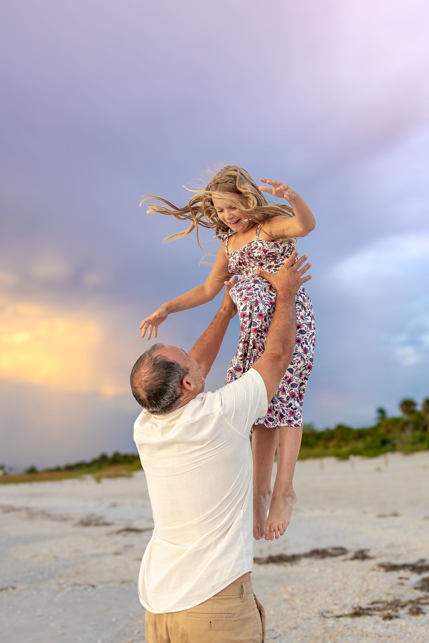 A man lifts a smiling girl in a floral dress on a sandy beach at sunset with dramatic purple clouds.