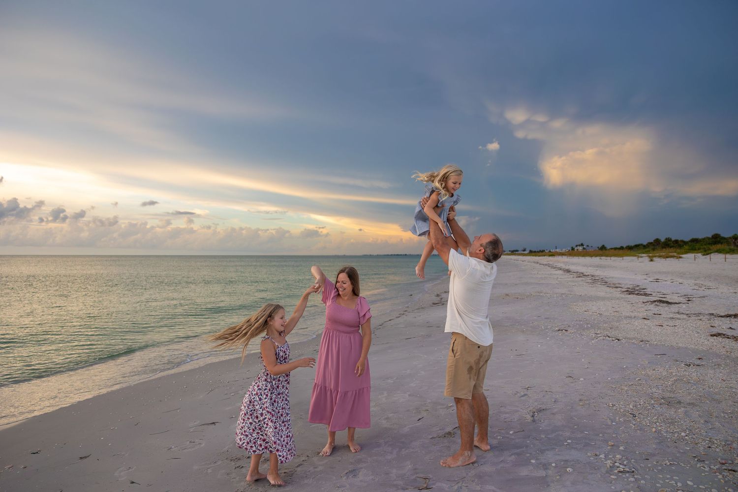 Family enjoying a sunset beach vacation, with father lifting baby in the air while mother and daughter watch joyfully.