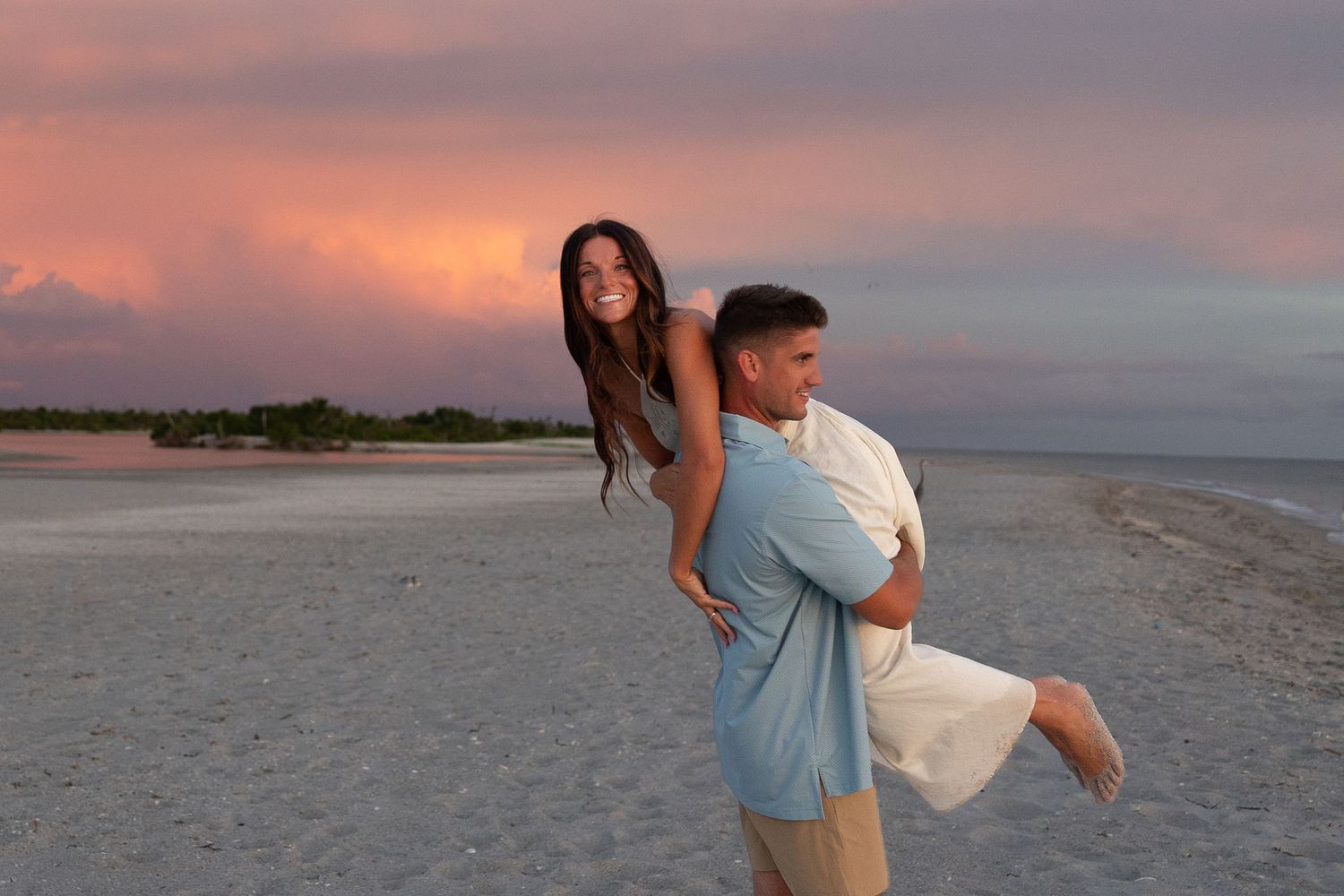 Couple enjoying a romantic piggyback ride on a sandy beach during a stunning pink and purple sunset.