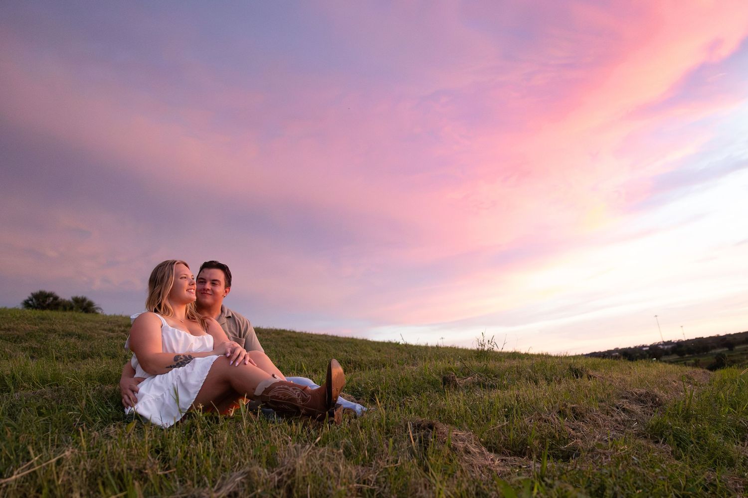 Couple sitting together on green hillside enjoying a stunning pink and purple sunset sky in a romantic outdoor setting.