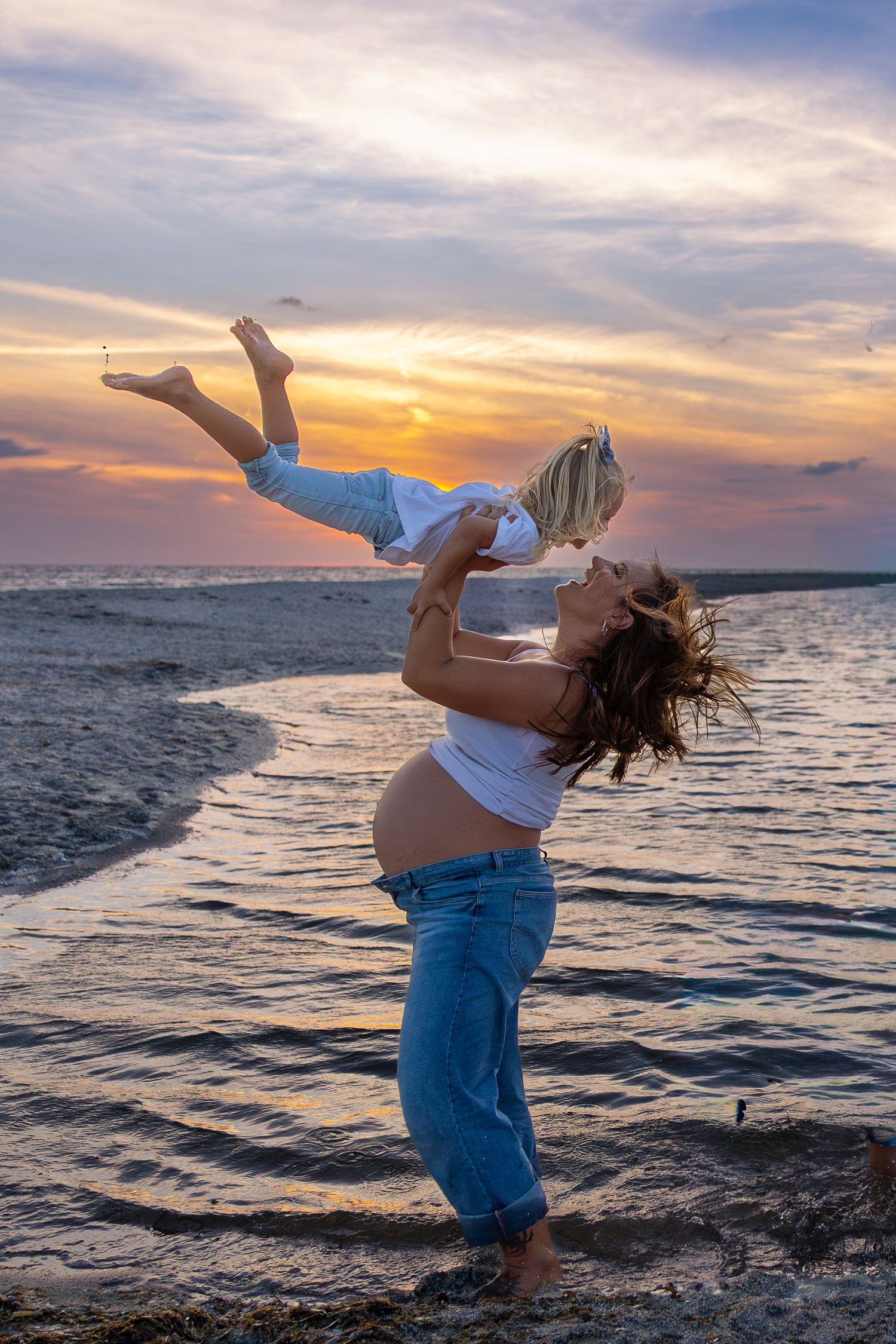 Pregnant woman lifting child at beach sunset, joyful maternity moment with colorful sky and ocean waves.