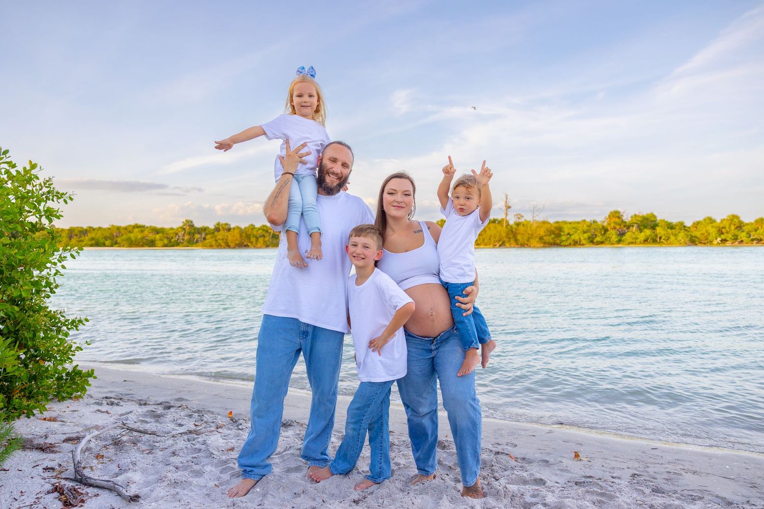 Family of five posing on a white sand beach with turquoise water and green trees in the background.