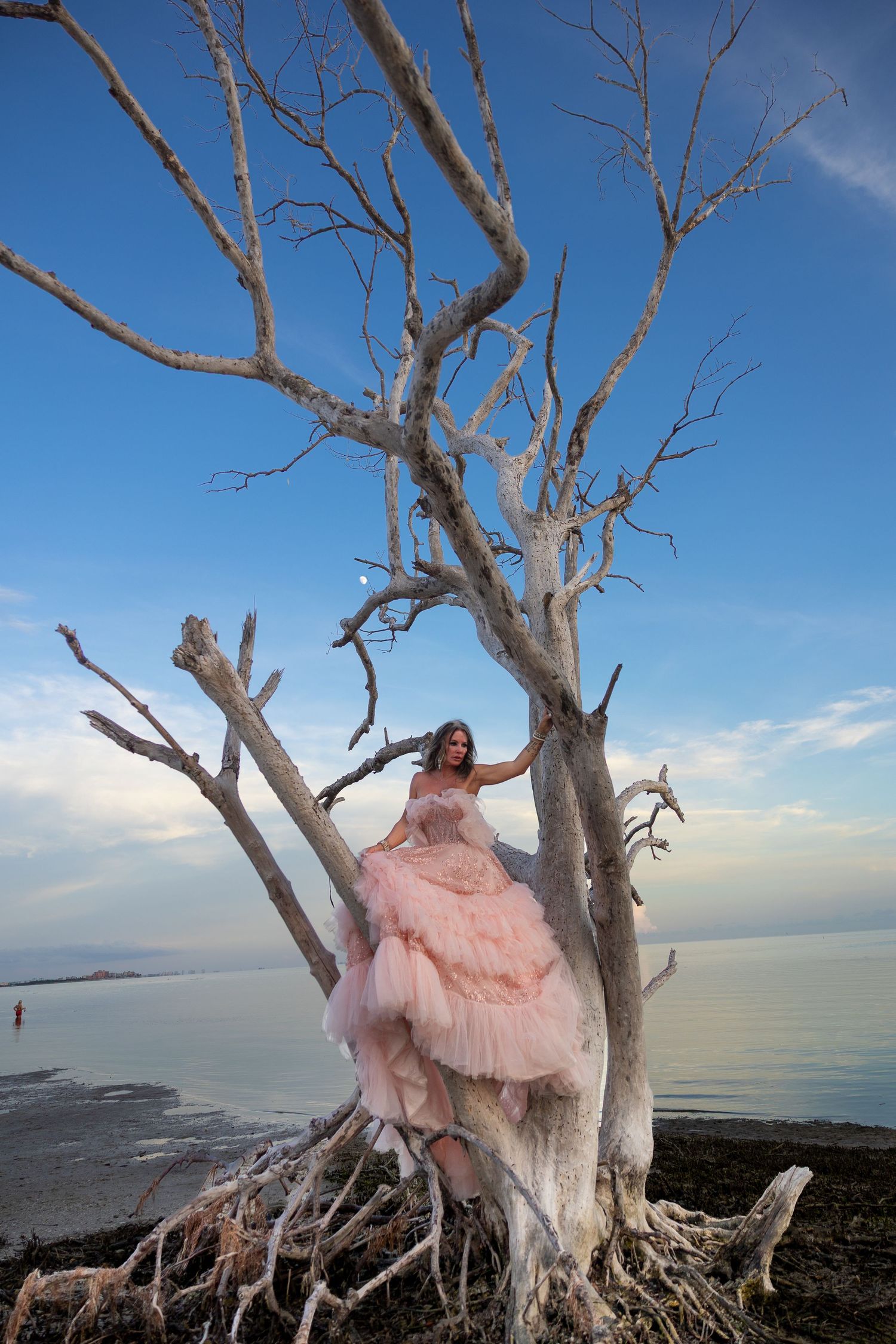 Woman in pink ball gown posing in bare white tree branches by the ocean at dusk.