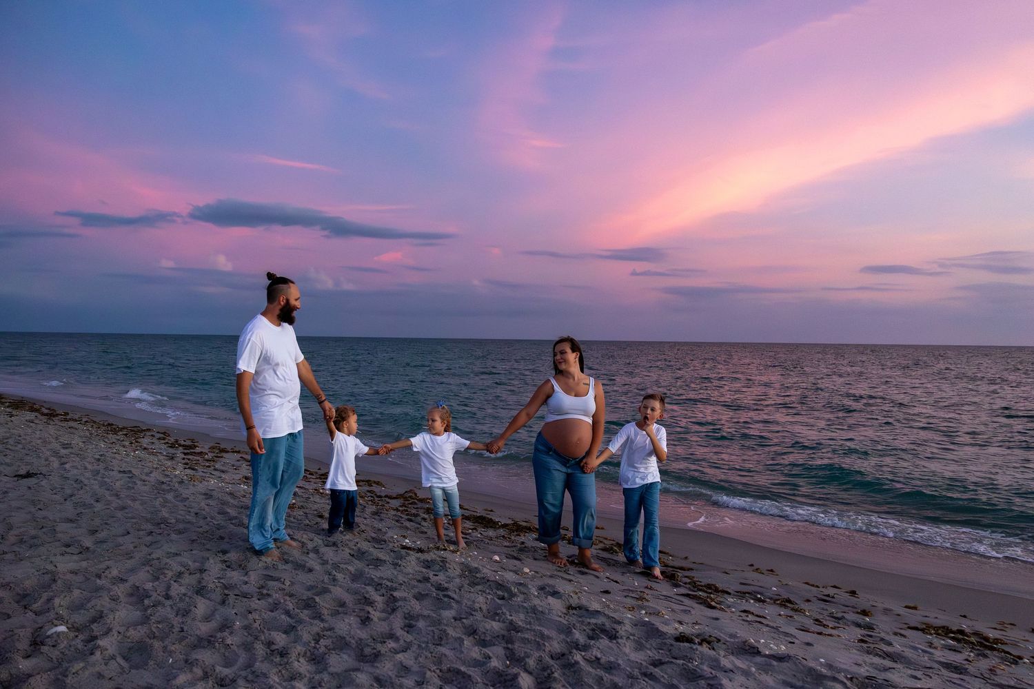 Pregnant family holding hands on beach at sunset with pink and purple sky.