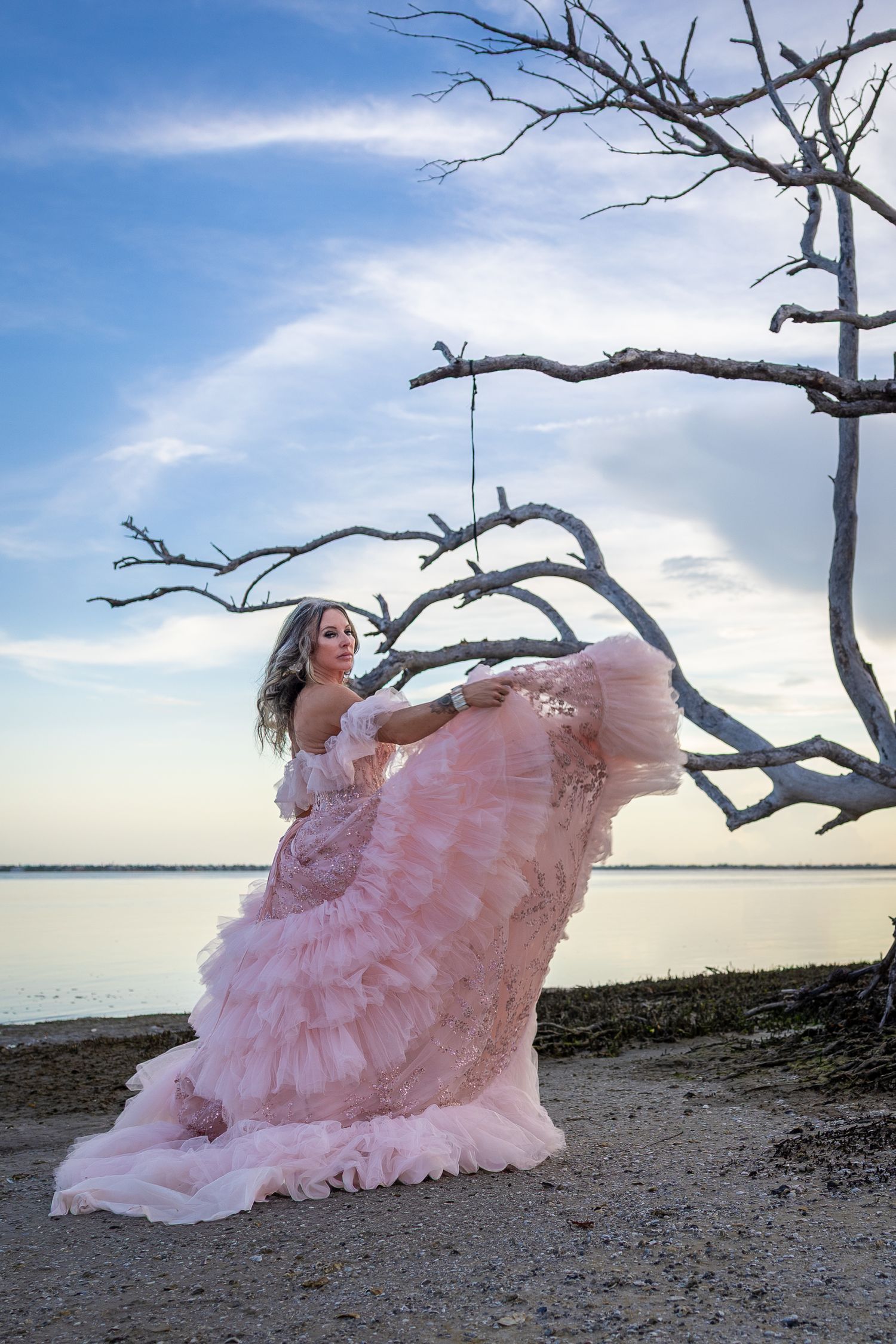 Woman in flowing pink ruffle gown poses near bare tree on sandy beach at sunset.