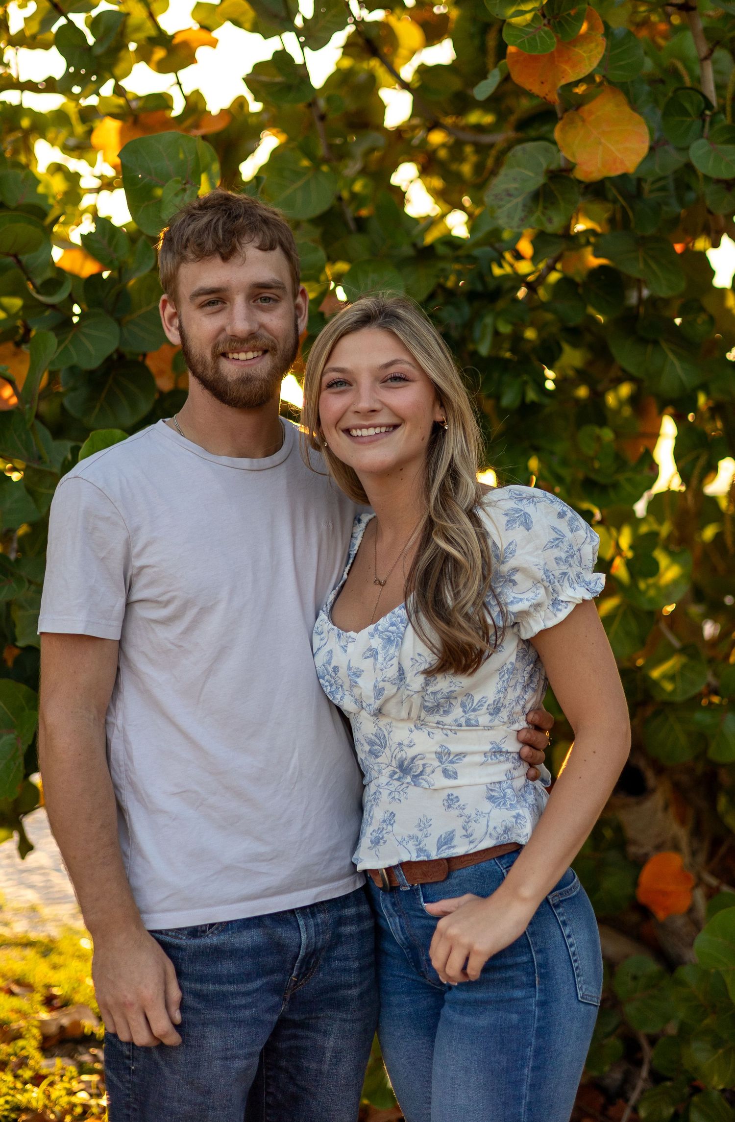 Smiling couple posing together outdoors in front of lush green trees with orange fruit in warm sunlight.