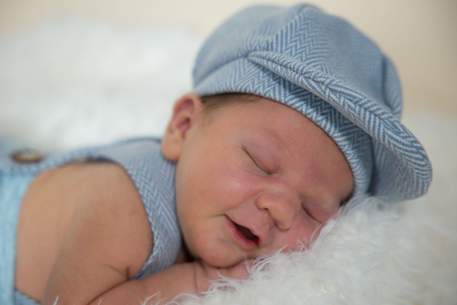 Sleeping newborn baby wearing a blue herringbone newsboy cap and matching outfit resting on a white fluffy blanket.