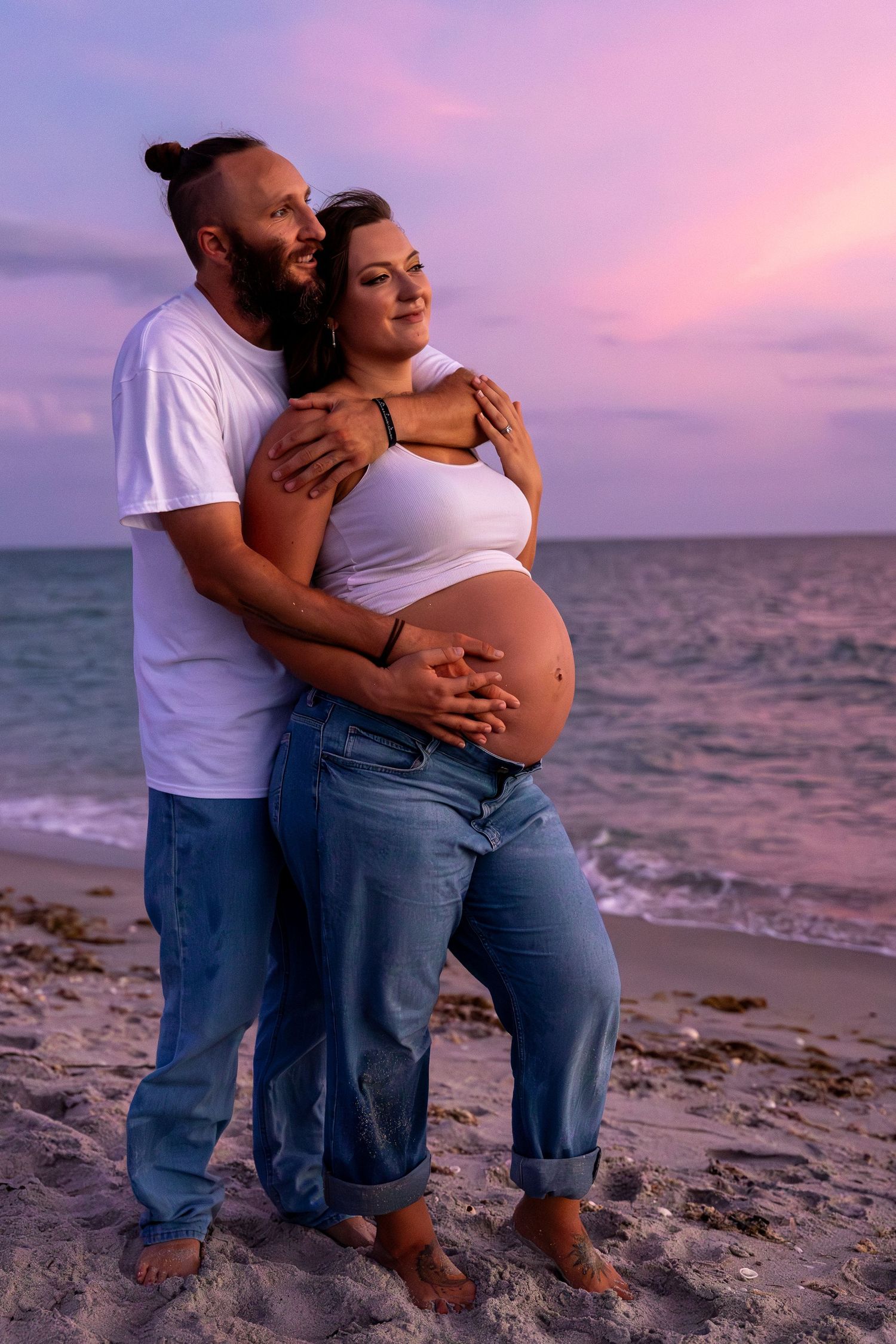 Couple embracing on beach at sunset, pregnant woman in white crop top and jeans with partner in white tee.