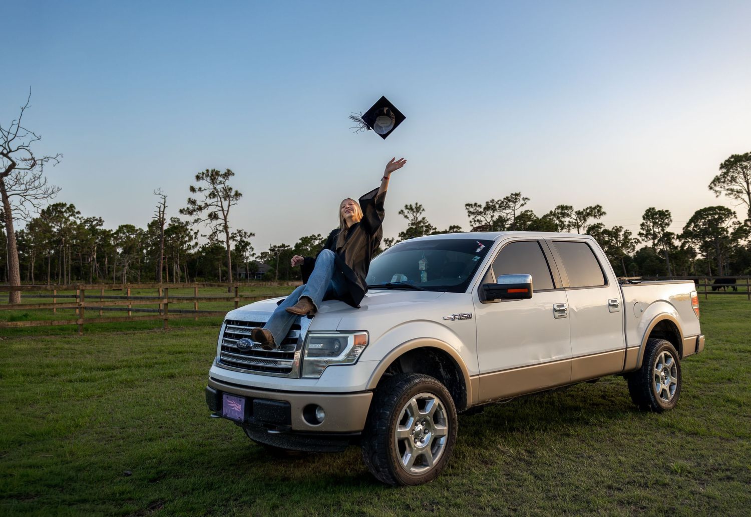 Graduate tosses cap from white pickup truck hood in scenic green field at dusk, celebrating academic achievement outdoors.