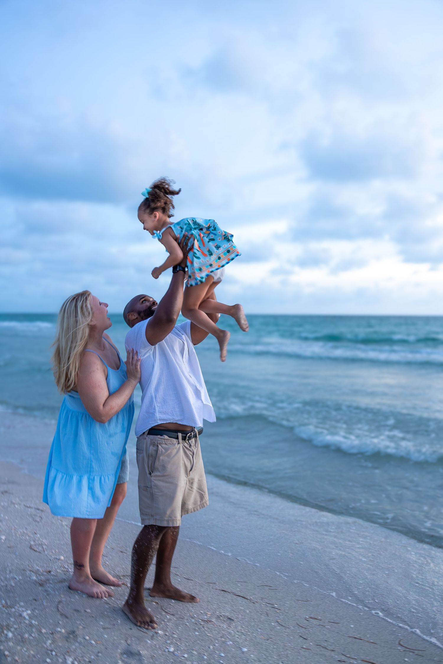 Family enjoying beach vacation, father lifting daughter in air while mother watches, ocean waves in background.