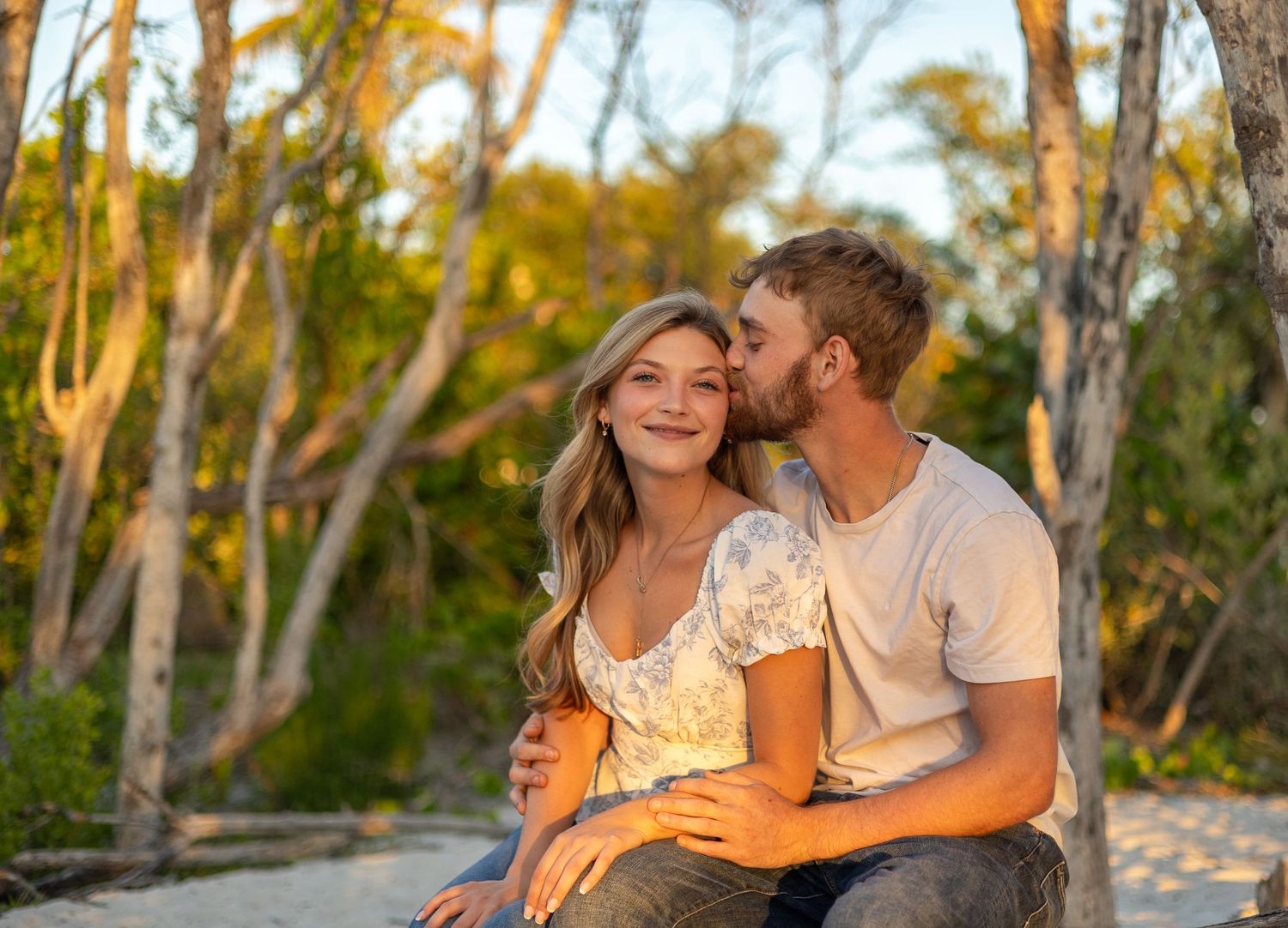 A romantic couple shares a tender kiss on a sandy beach with sun-lit trees in the background.