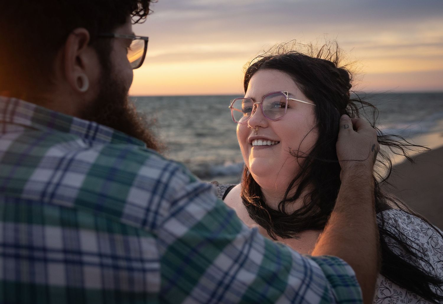 Couple sharing a romantic moment at sunset on the beach, woman smiling as man brushes hair from her face.