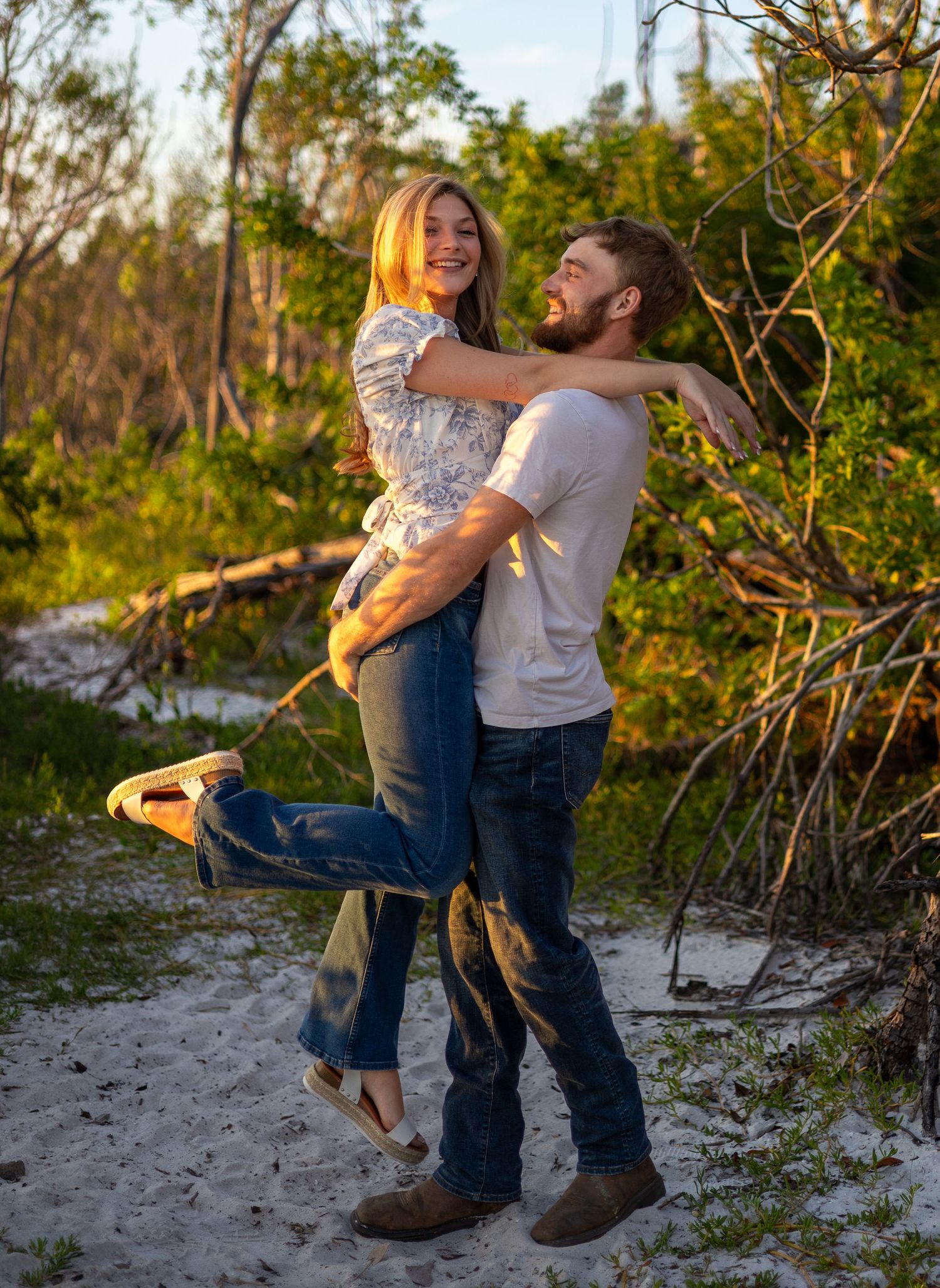 A joyful man lifts a laughing woman in his arms on a sandy path surrounded by lush green trees.