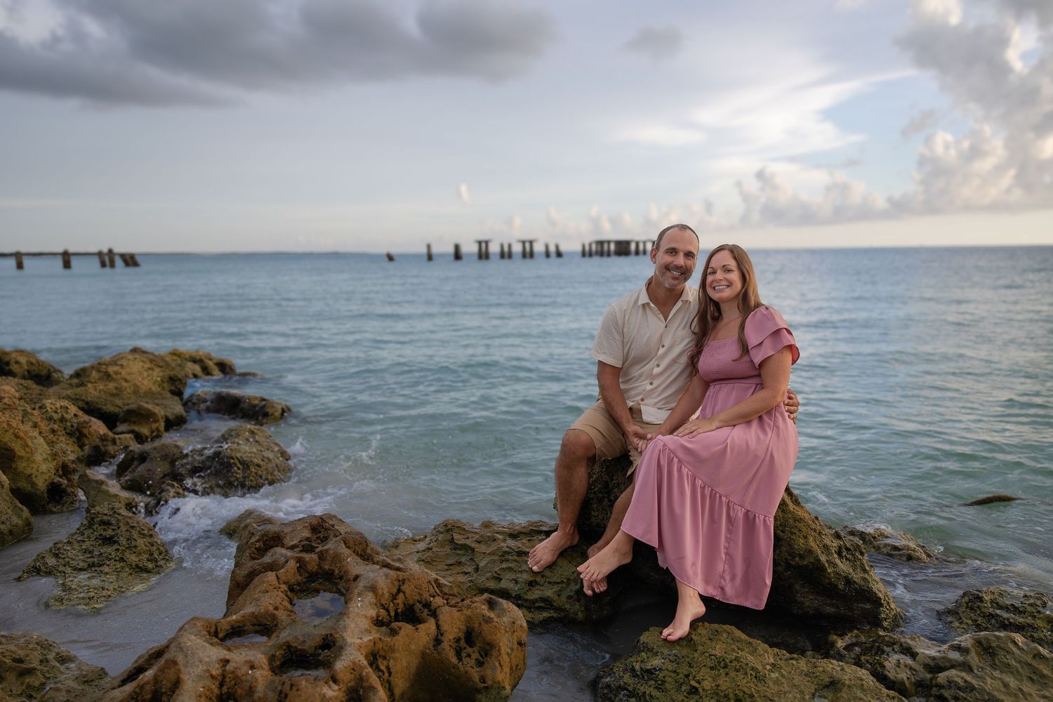 Couple sitting on rocky shoreline by turquoise ocean at sunset, woman in pink dress, man in white shirt.