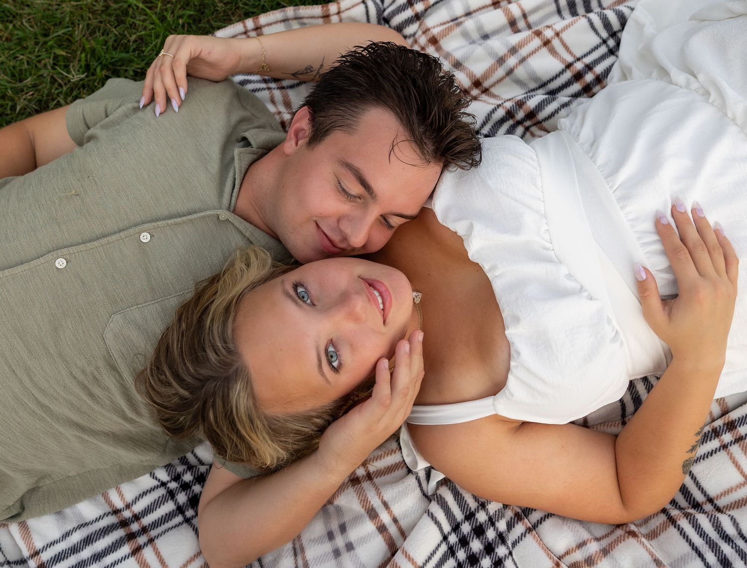 A couple lying on a plaid blanket outdoors, sharing a tender romantic moment while cuddling together.