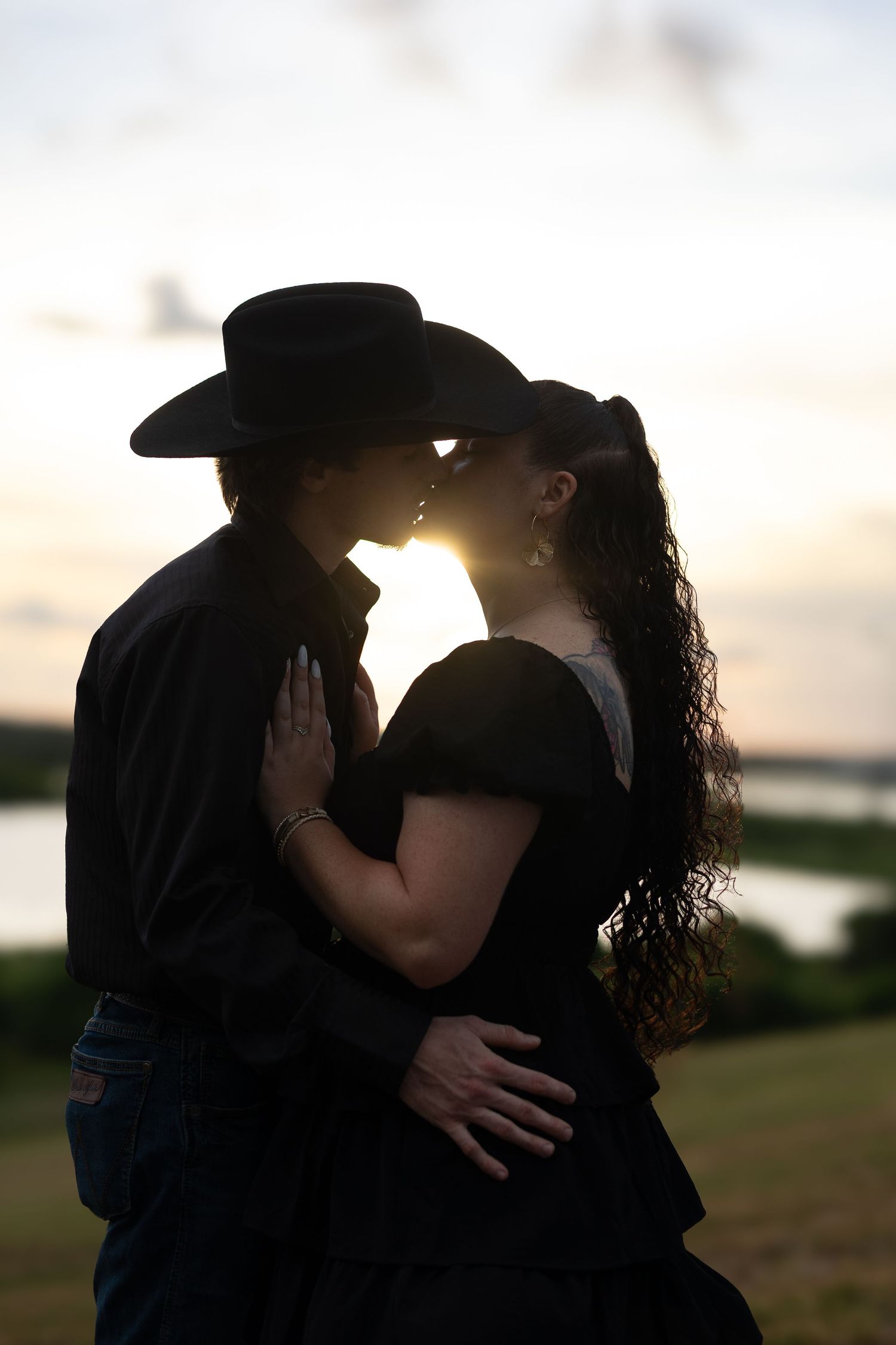Silhouette of a couple kissing at sunset, man in cowboy hat embracing woman with long curly hair near a lake.