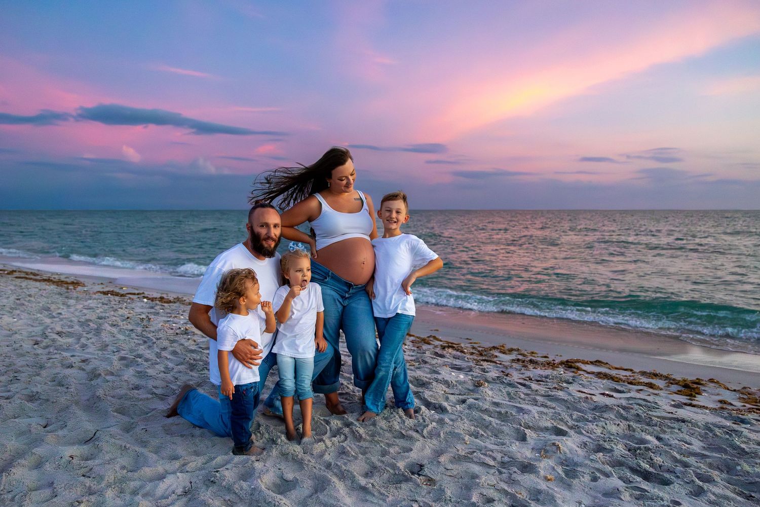 Pregnant woman with family on beach at sunset, wearing white shirts and jeans, posing together by the ocean.
