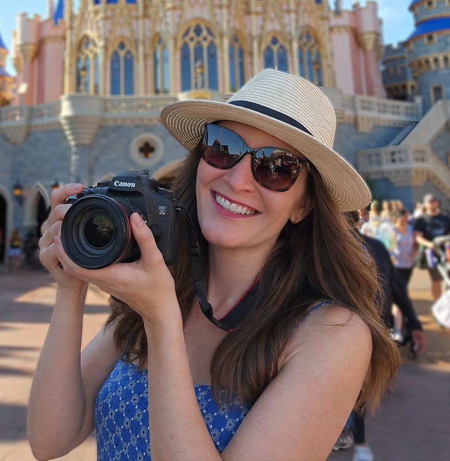 Happy woman wearing sunglasses and a straw hat photographs with a Canon camera at a Disney theme park.