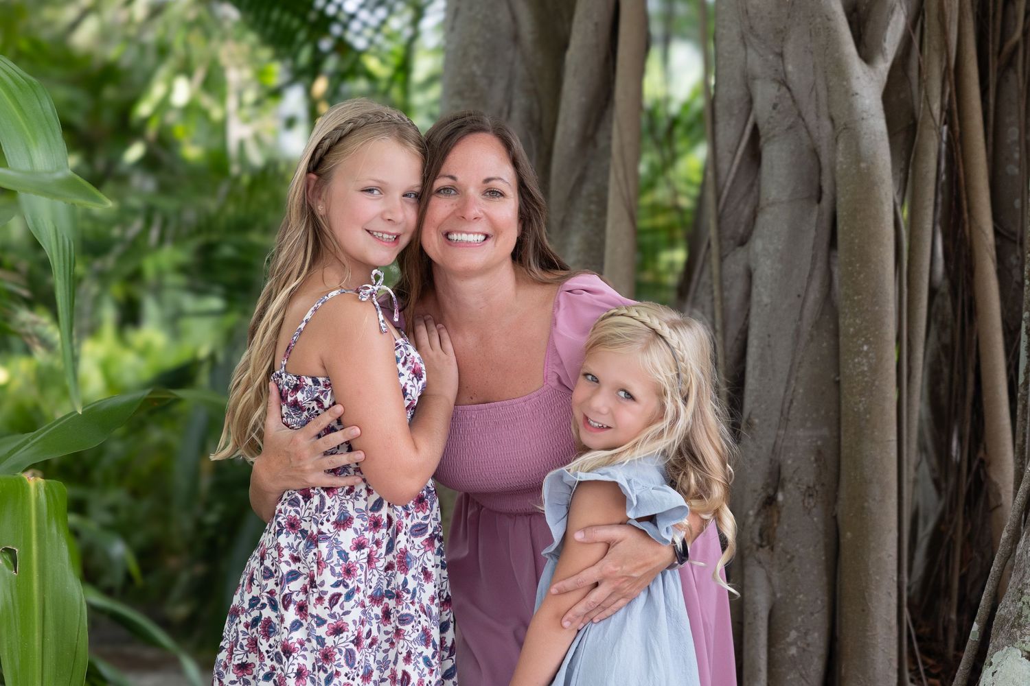 A smiling woman hugs two young blonde girls outdoors near a large tropical tree with lush green foliage.