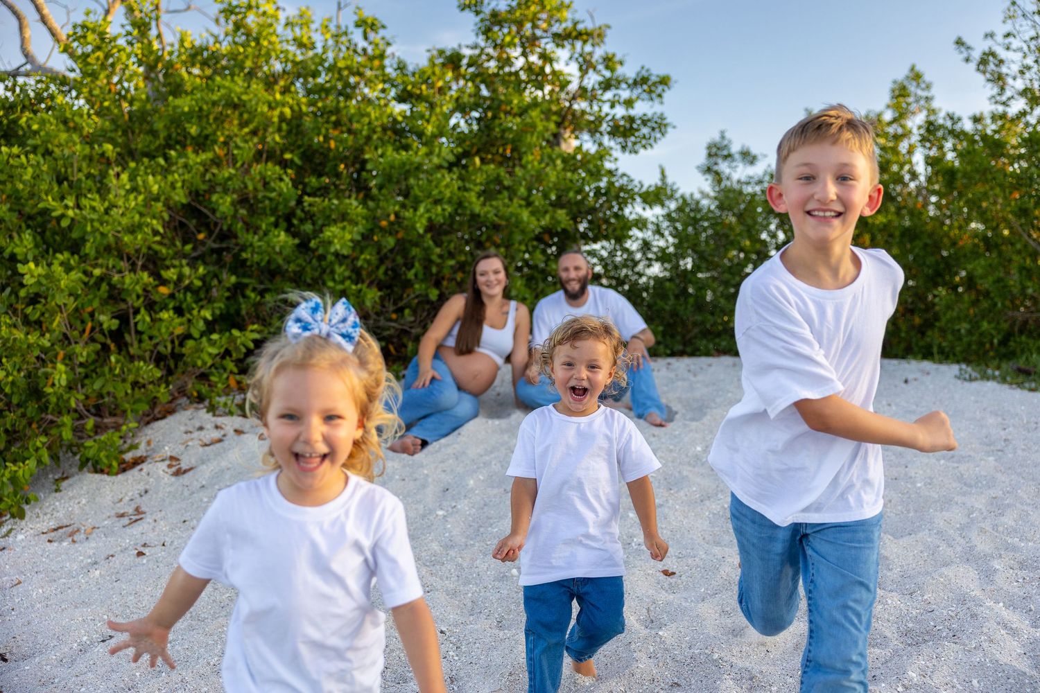 Three happy children run toward camera on sandy beach while parents sit together in background.