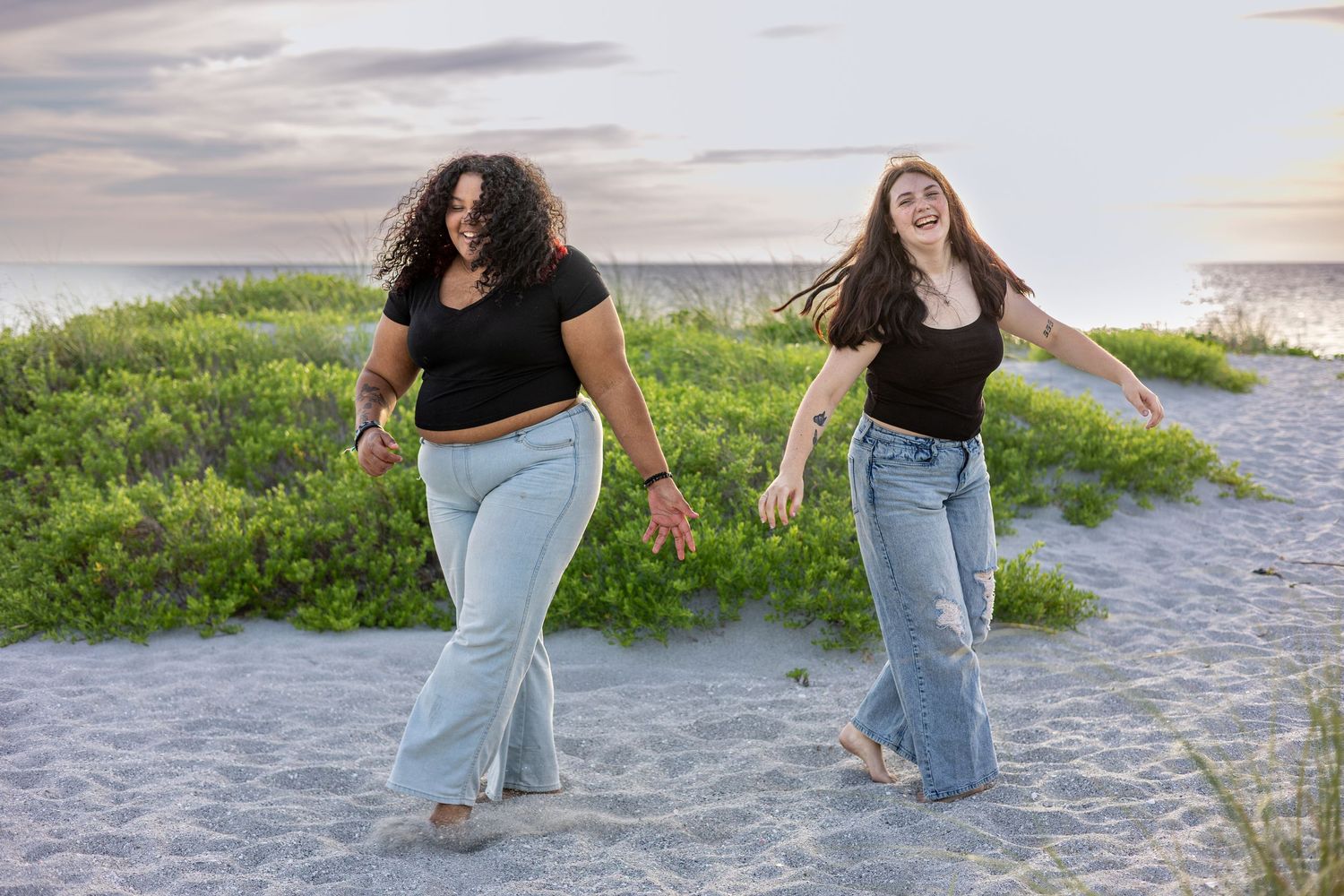Two women in black tops and jeans walking and laughing on sandy beach dunes.