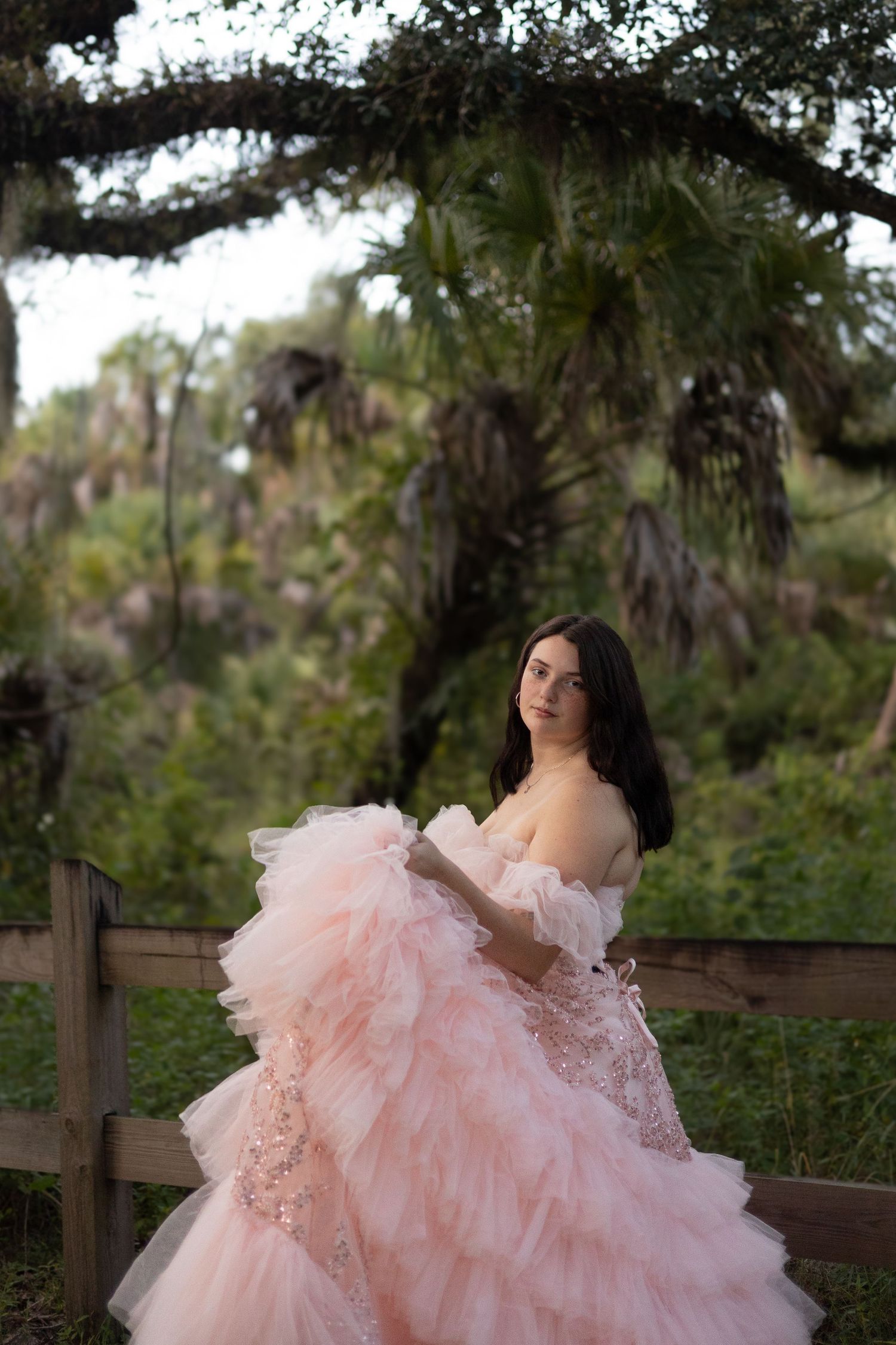 A young woman in a voluminous blush pink tulle ball gown poses gracefully outdoors near a wooden fence and lush greenery.