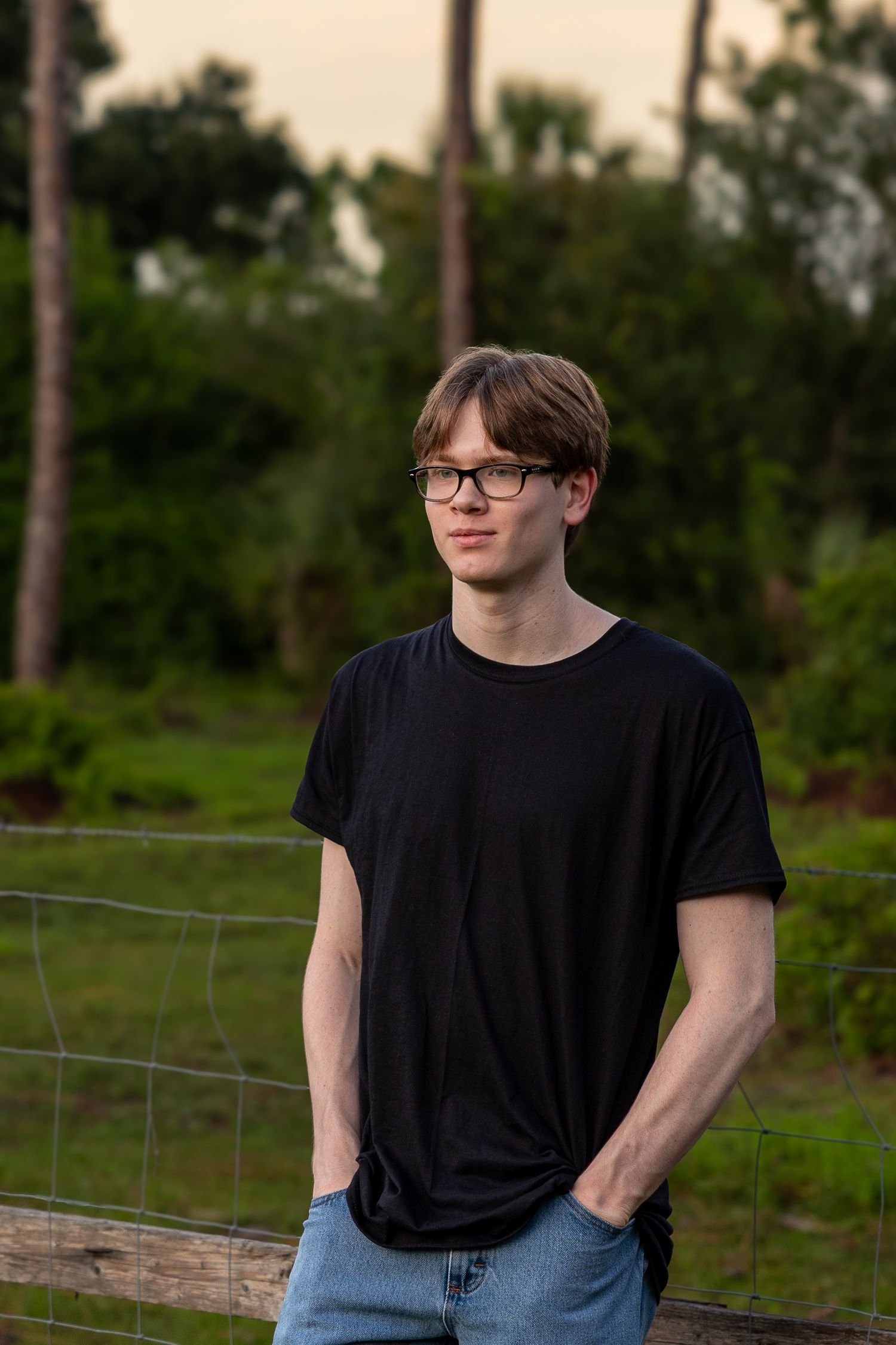 Young man in black t-shirt and glasses standing outdoors near a fence with green trees in background.