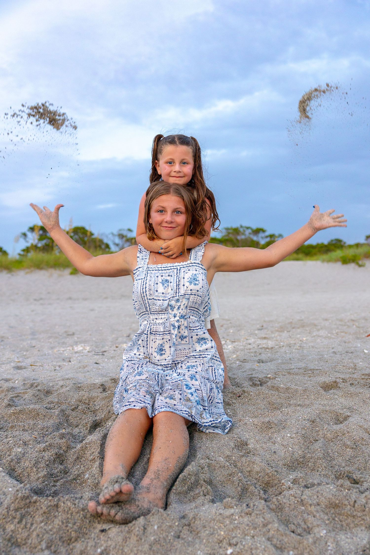 Two girls playing on a sandy beach, tossing sand in the air, wearing summer dresses with sandy feet.
