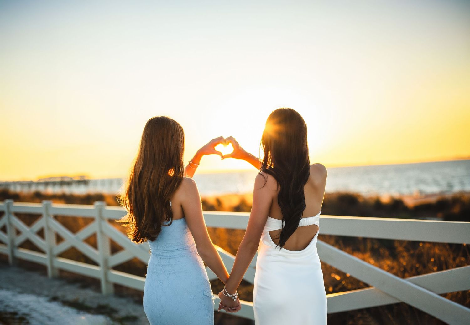 Two women in white dresses making a heart shape with hands at beach sunset.