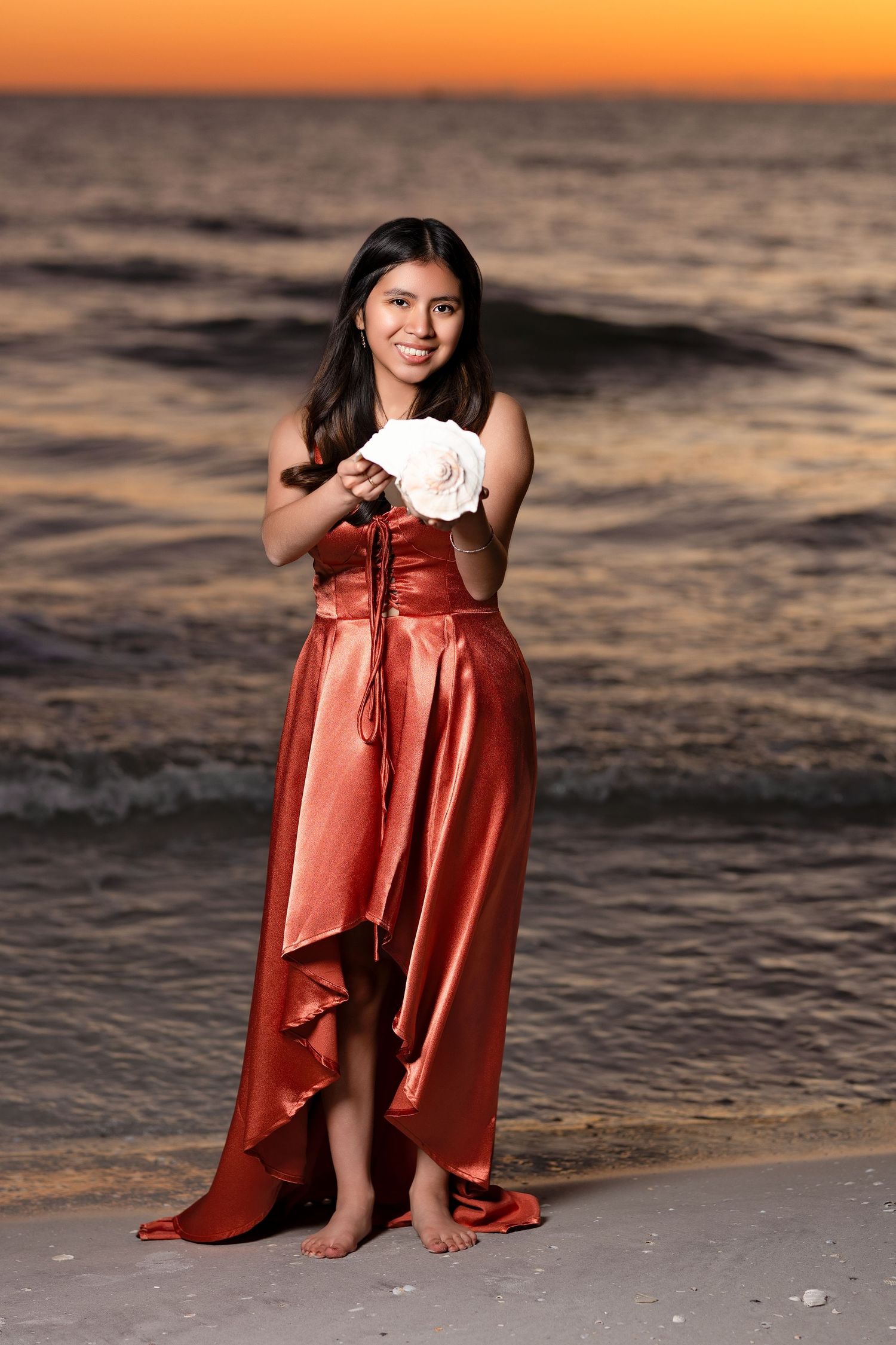 Young woman in red satin gown holding white roses posing on beach at vivid sunset.
