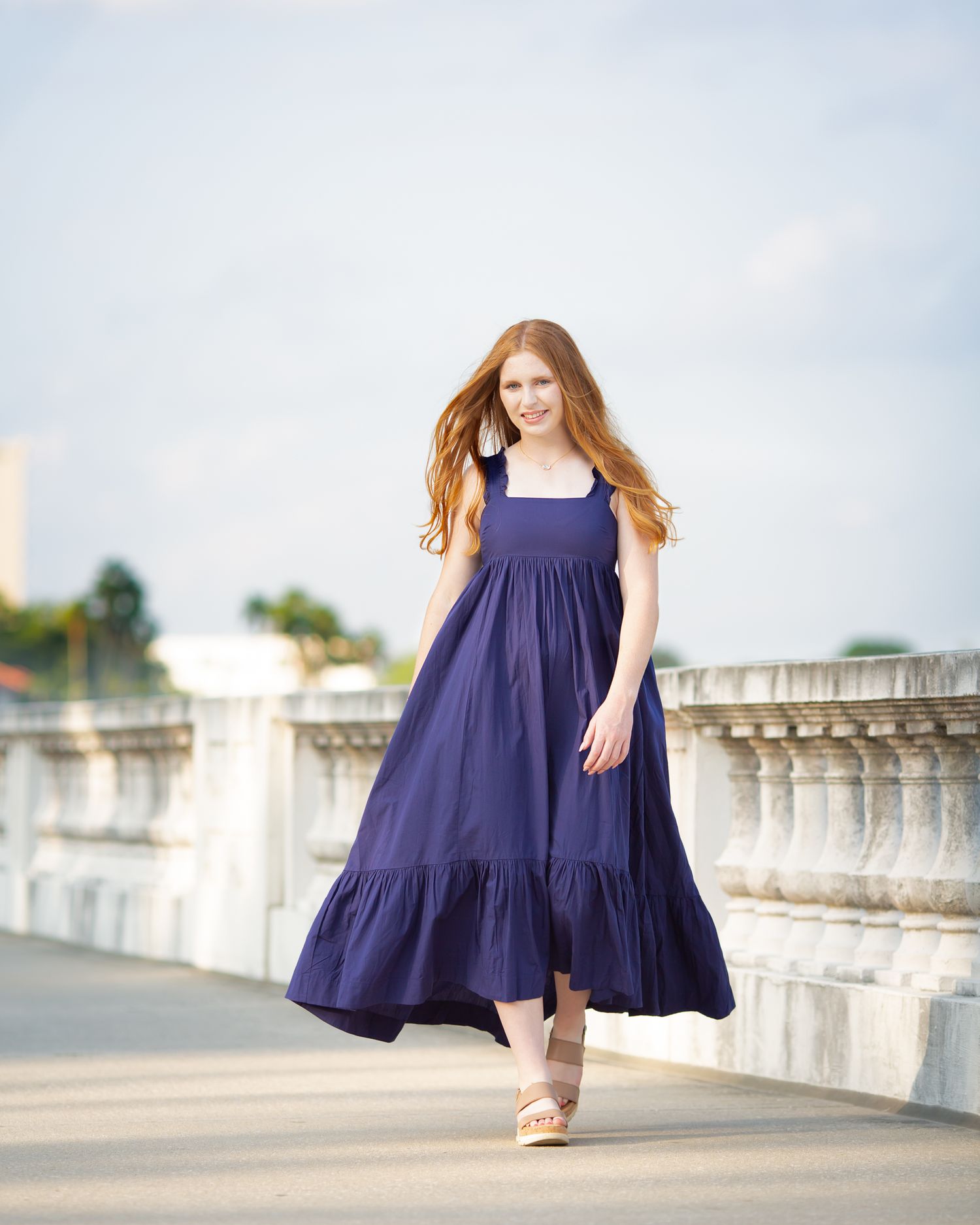 Redhead woman in navy blue maxi dress walking on outdoor bridge terrace.