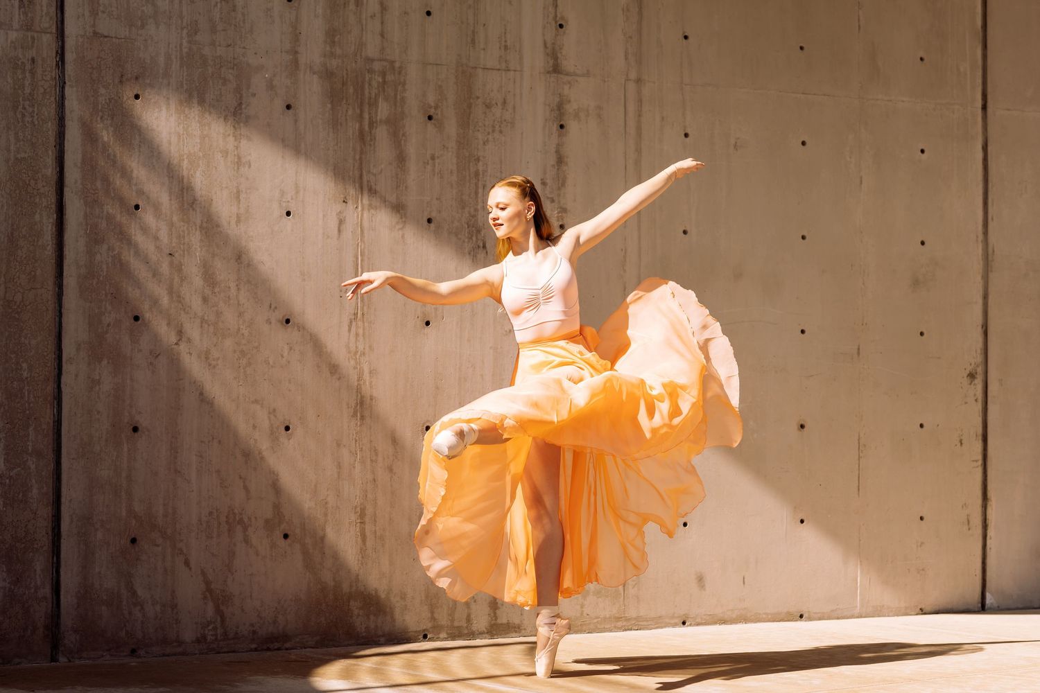 Graceful dancer in orange flowing skirt performing ballet pose in sunlit concrete space.