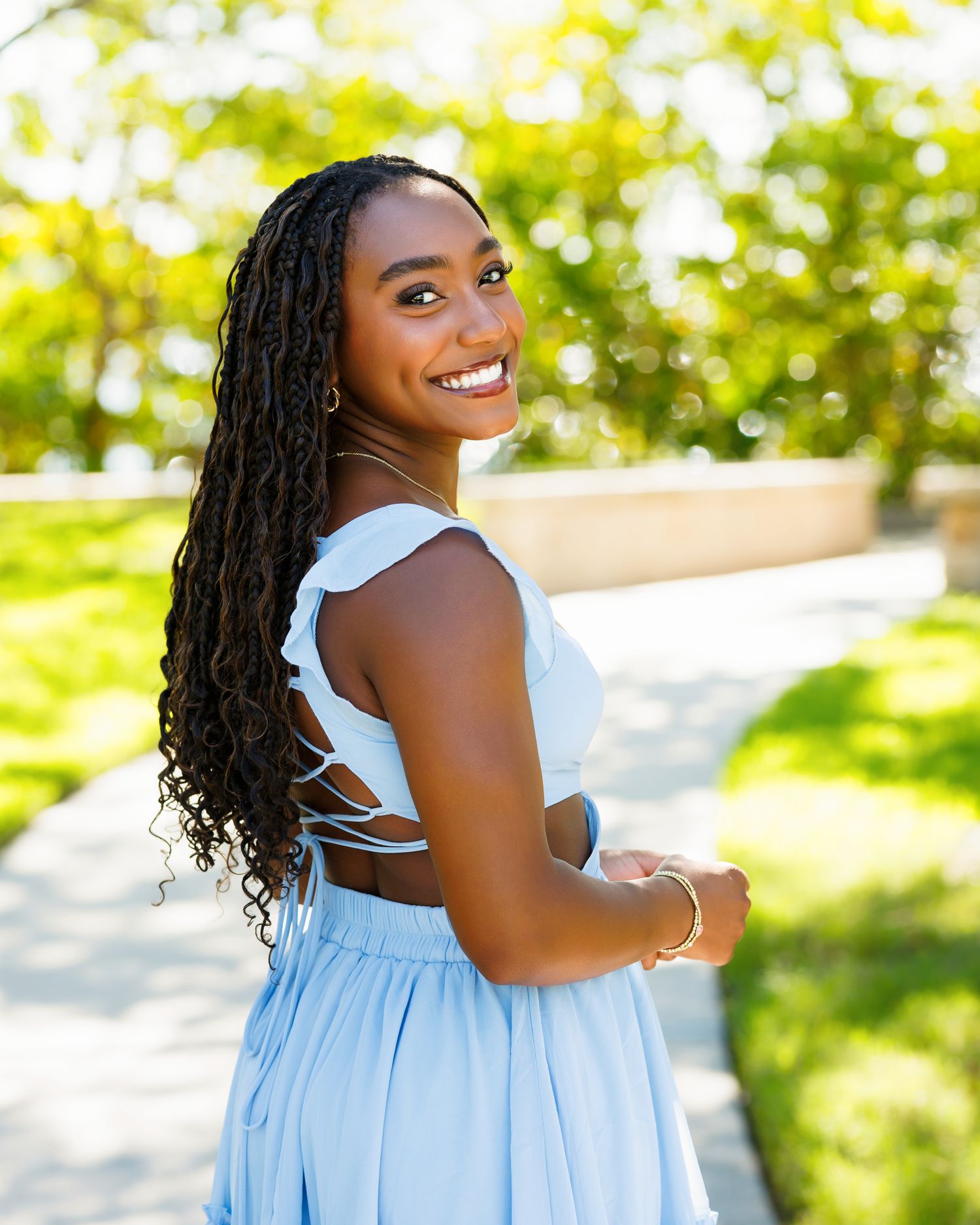 Smiling young Black woman with long braids wearing a light blue dress poses outdoors in golden sunlight.