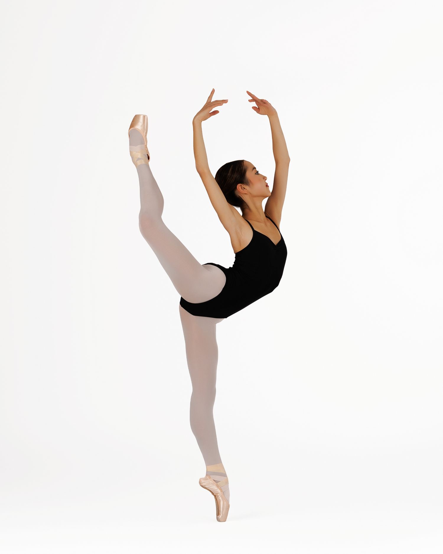 Ballet dancer in black leotard performs arabesque on pointe shoes against white background.