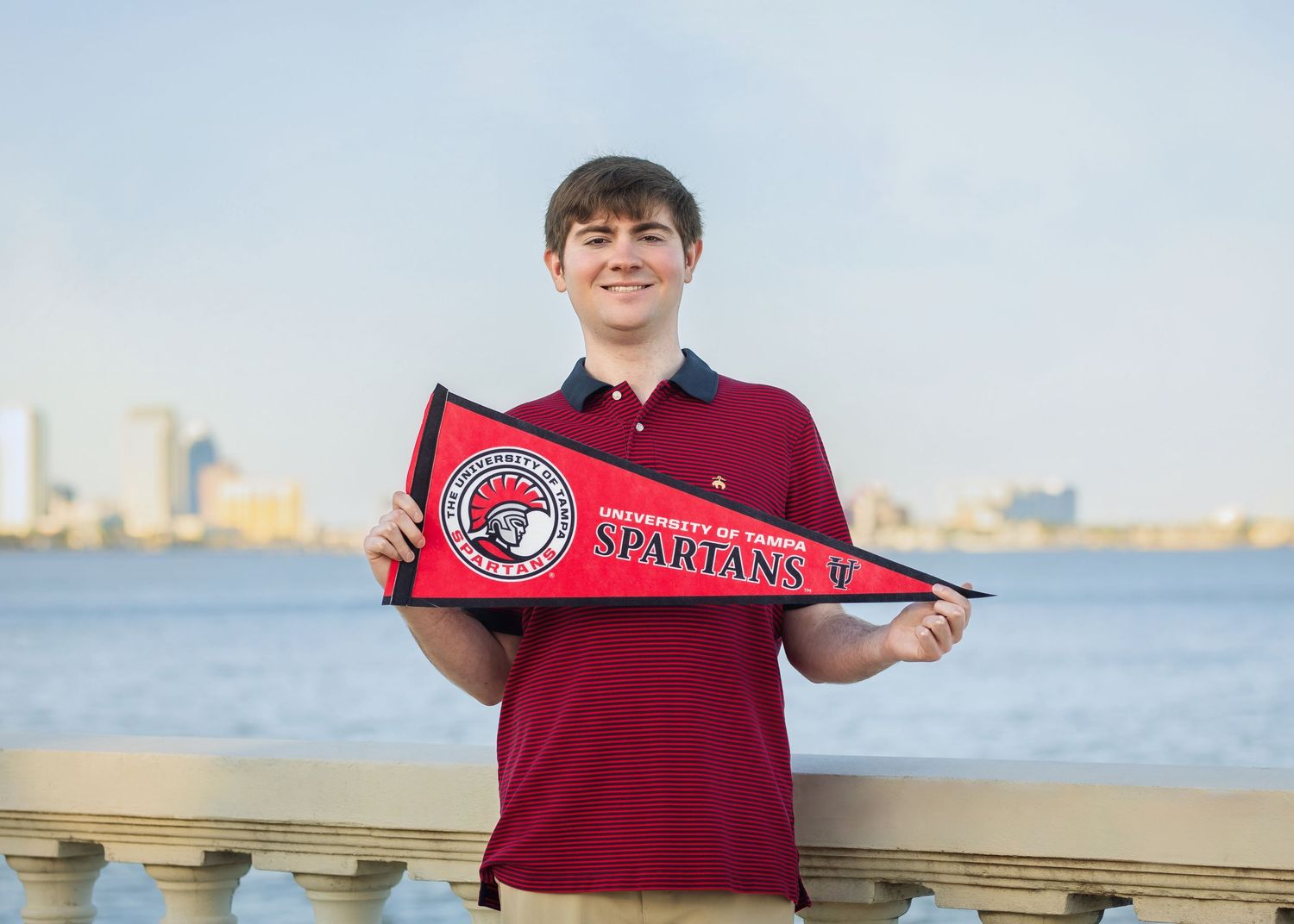 Smiling young man holding red Spartans pennant with city skyline and river in background.