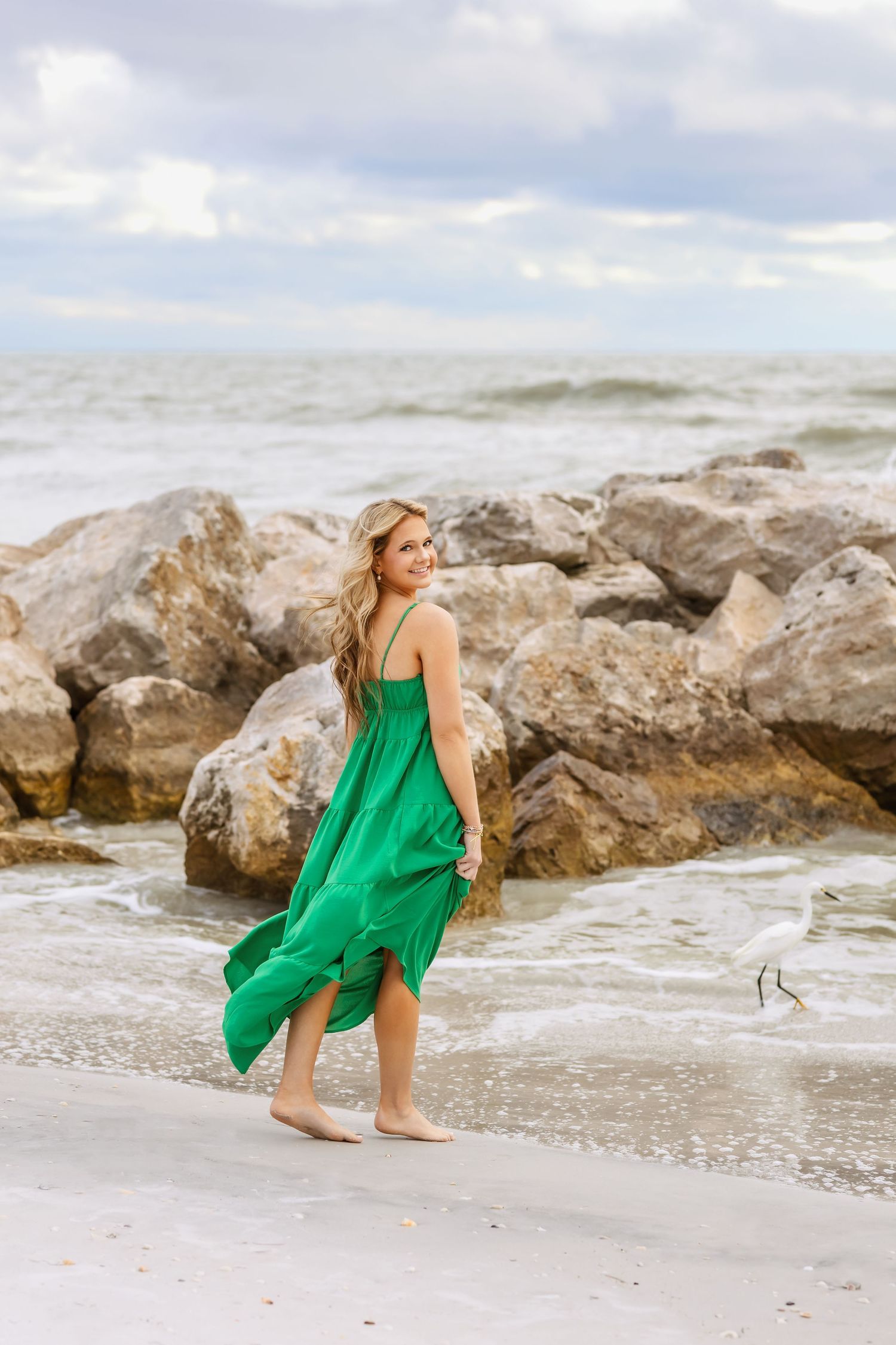 Blonde woman in flowing green dress walking barefoot on sandy beach near rocky shoreline.