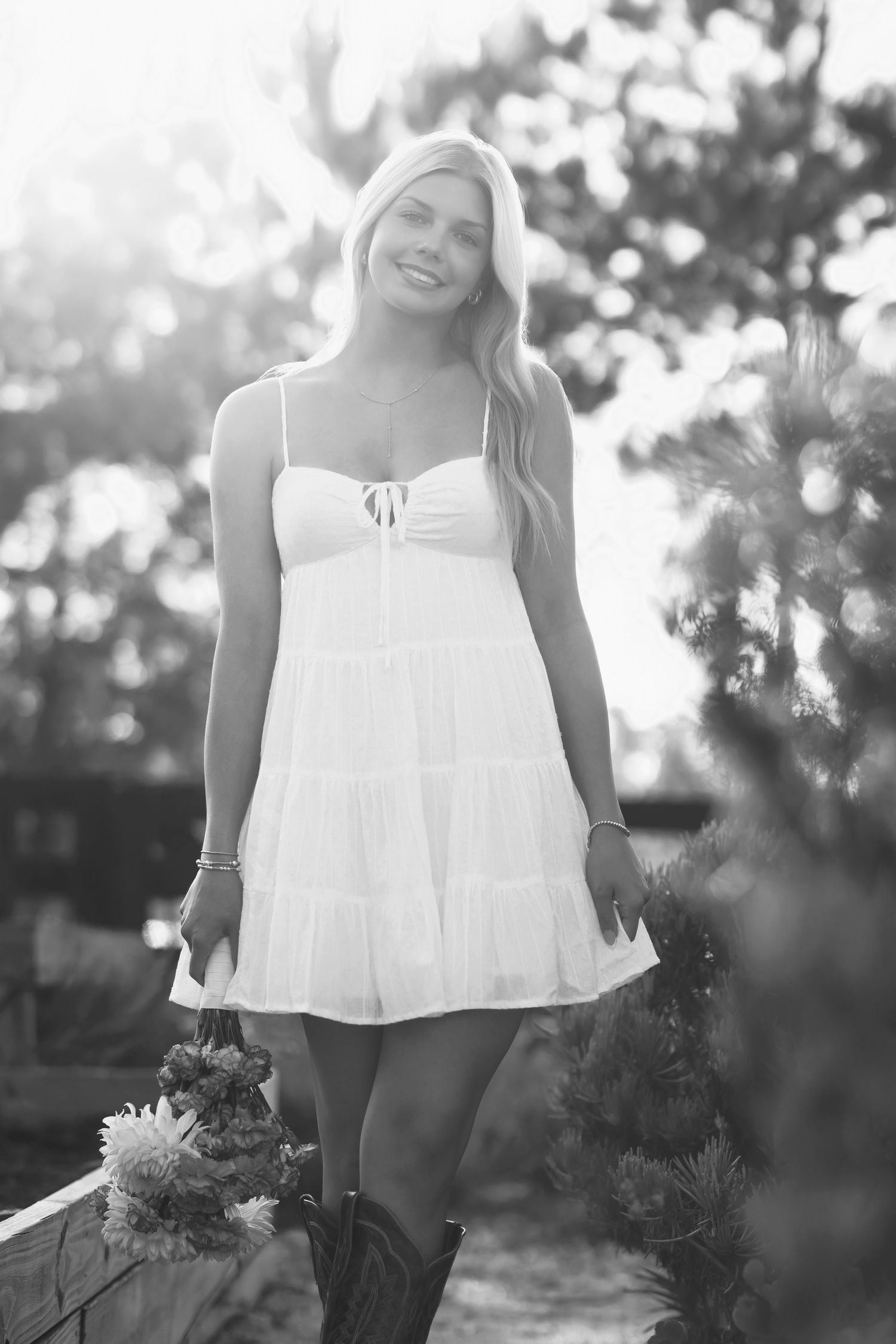 Smiling blonde woman in white sundress and cowboy boots posing outdoors in black and white photo.