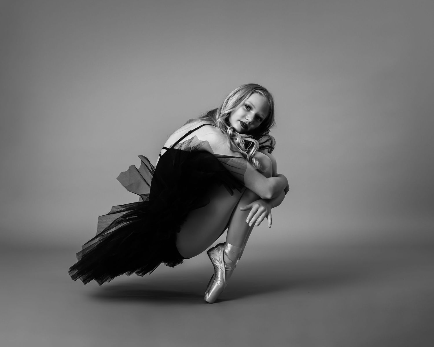Young ballerina in black tutu sitting in curled pose wearing pointe shoes, black and white.